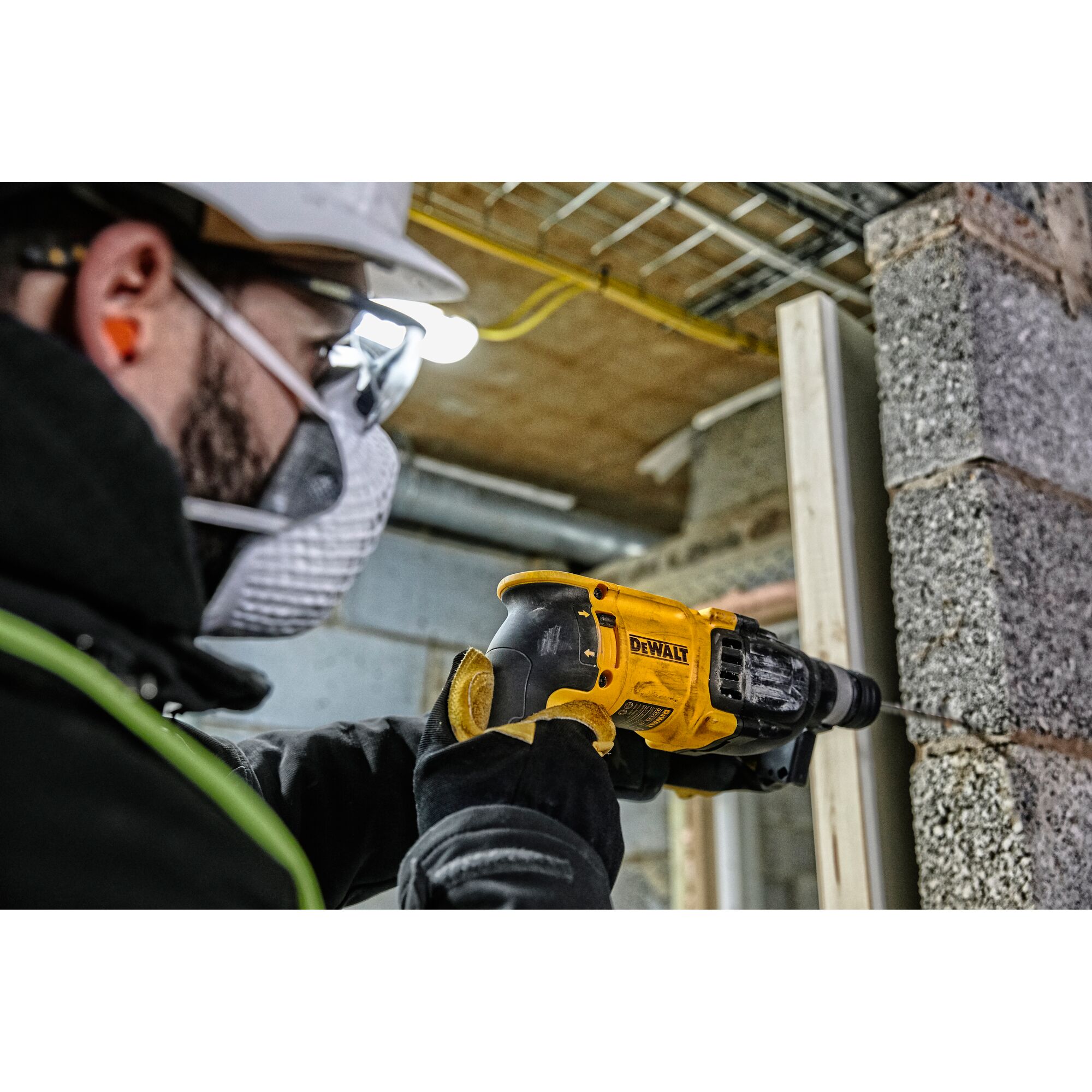 A person wearing safety gear uses a yellow DEWALT D25133K rotary hammer drill to work on a concrete wall in a construction setting.