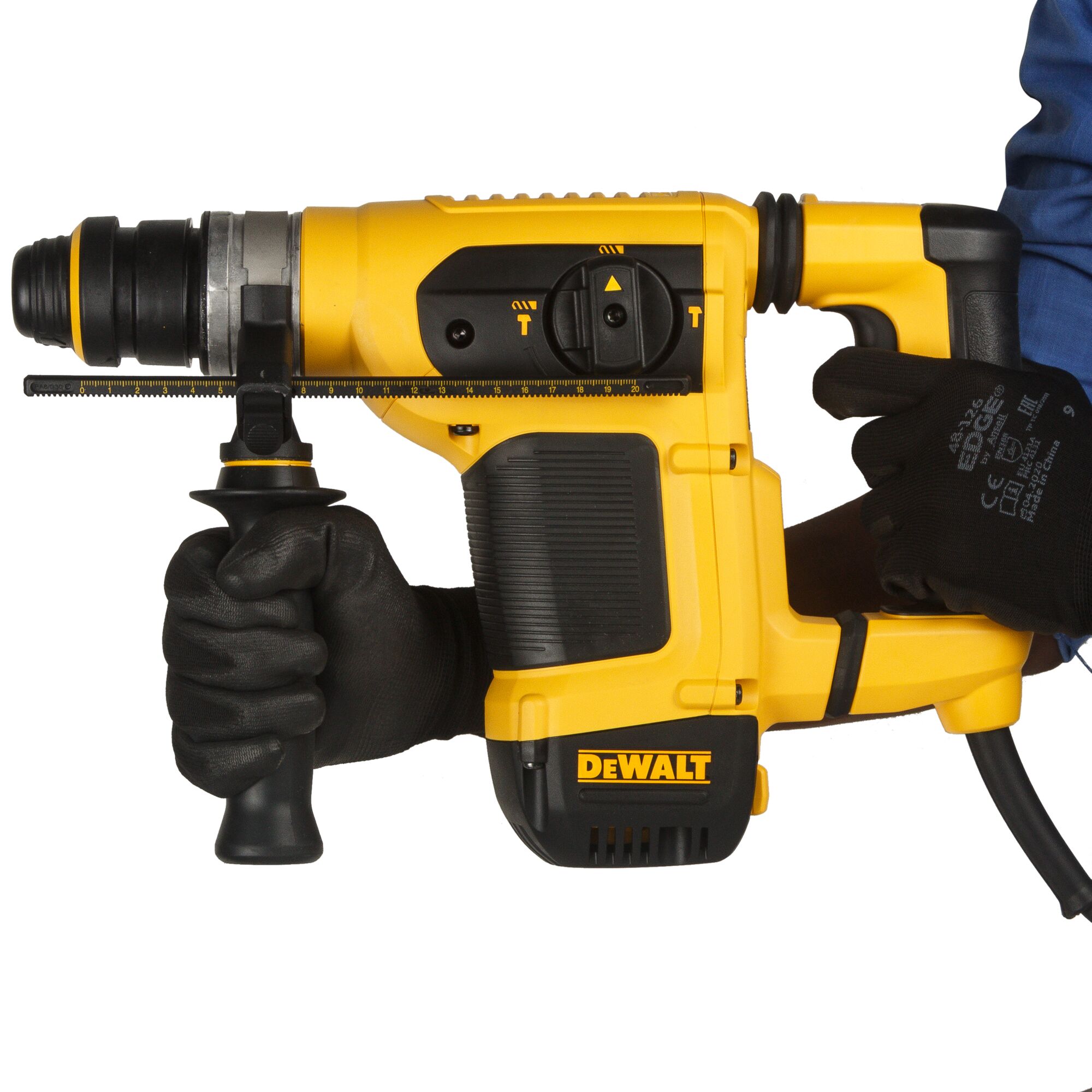 A person wearing black gloves holds a yellow DEWALT rotary hammer drill, model D25413K, designed for heavy-duty drilling and construction tasks.