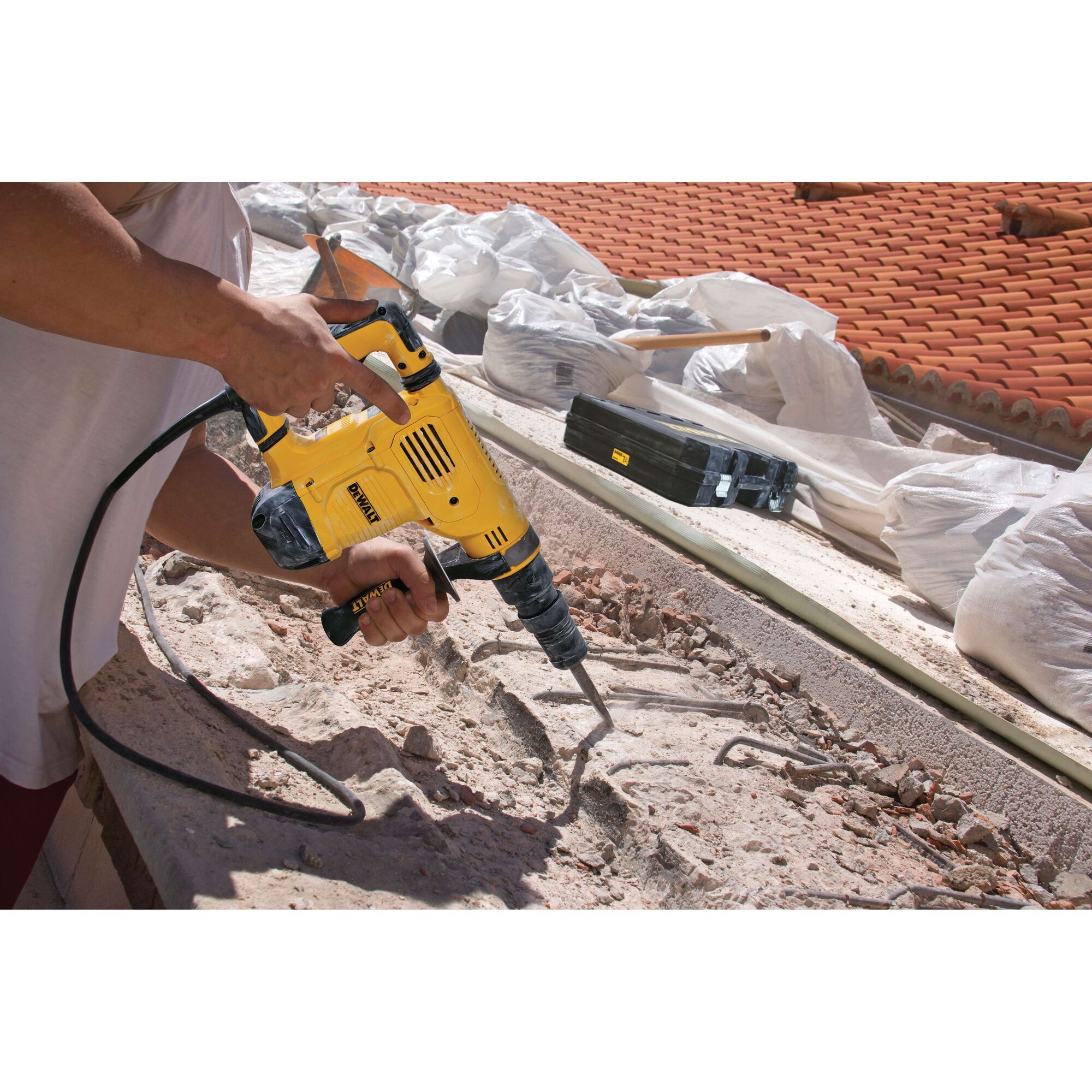 A person using a yellow DEWALT electric demolition hammer to break up concrete and rubble outdoors, with construction materials and tiled roofing visible in the background.