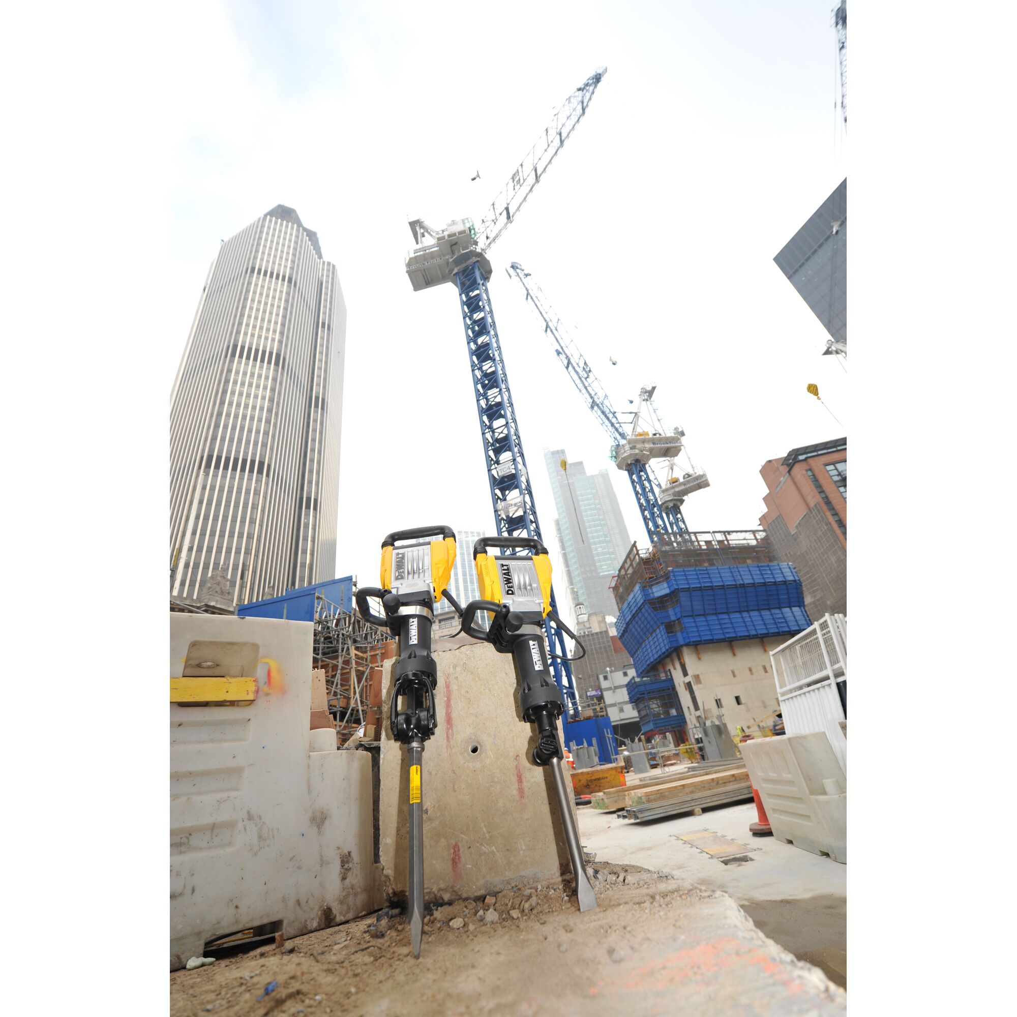 Two DEWALT demolition hammers (model D25961K) with yellow and black handles are standing upright at a construction site. In the background, there are tall modern buildings and several construction cranes extending into the sky.