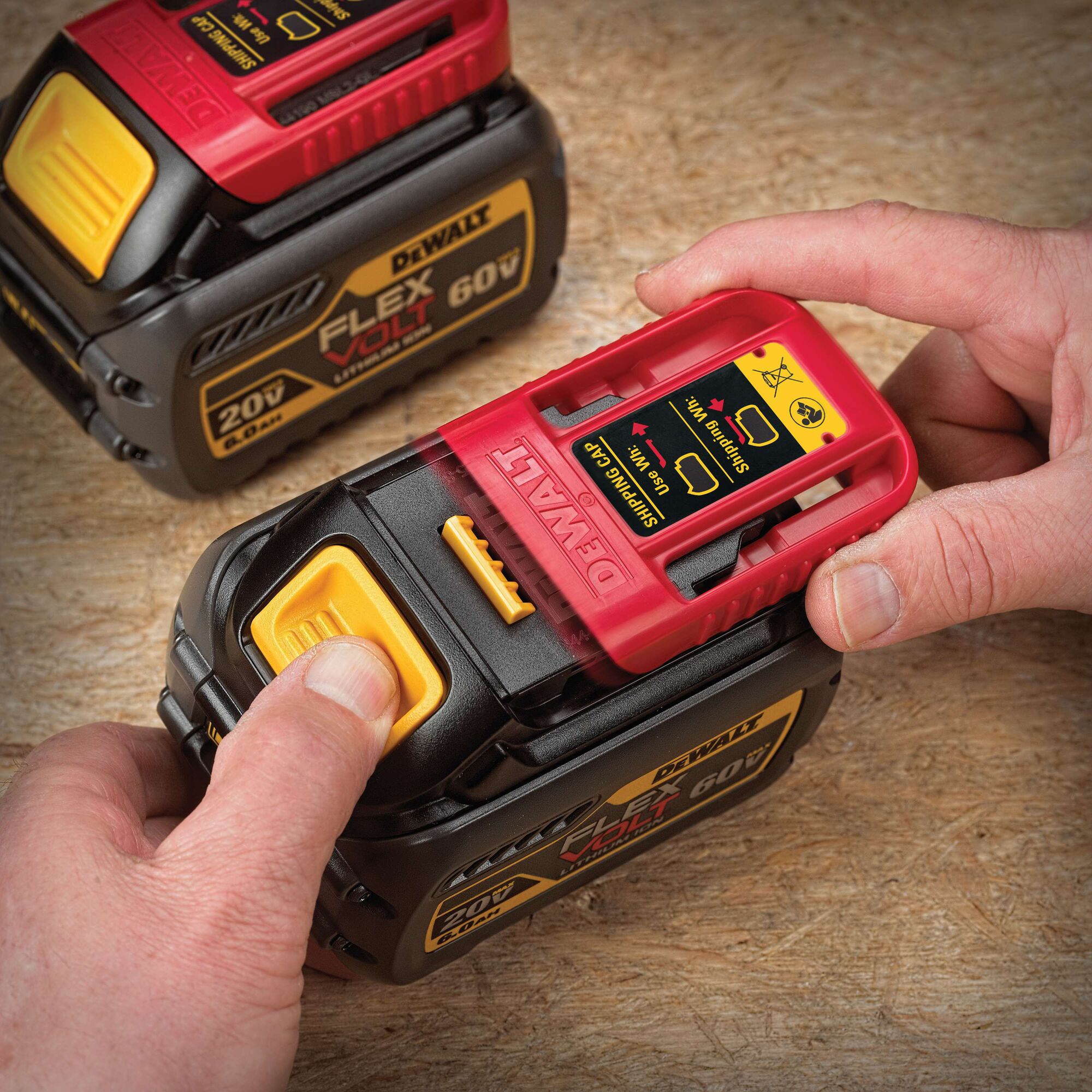 A close-up of hands operating a DEWALT FlexVolt 60V/20V rechargeable battery pack on a wooden surface. One battery has a red shipping cap being removed, showing safety instructions. The product branding and voltage details are clearly visible.