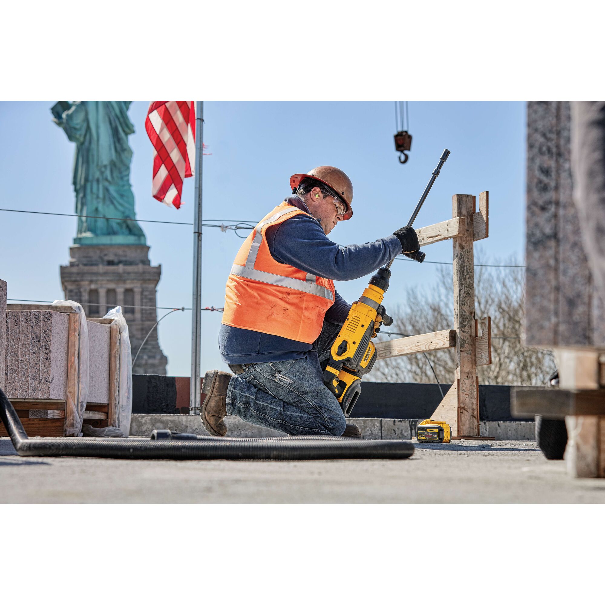 A construction worker wearing an orange safety vest and helmet uses a yellow DEWALT power tool for drilling at a construction site. The worker is kneeling near wooden structures, with the DEWALT DCB612 battery visible beside them. The Statue of Liberty and an American flag are visible in the background.