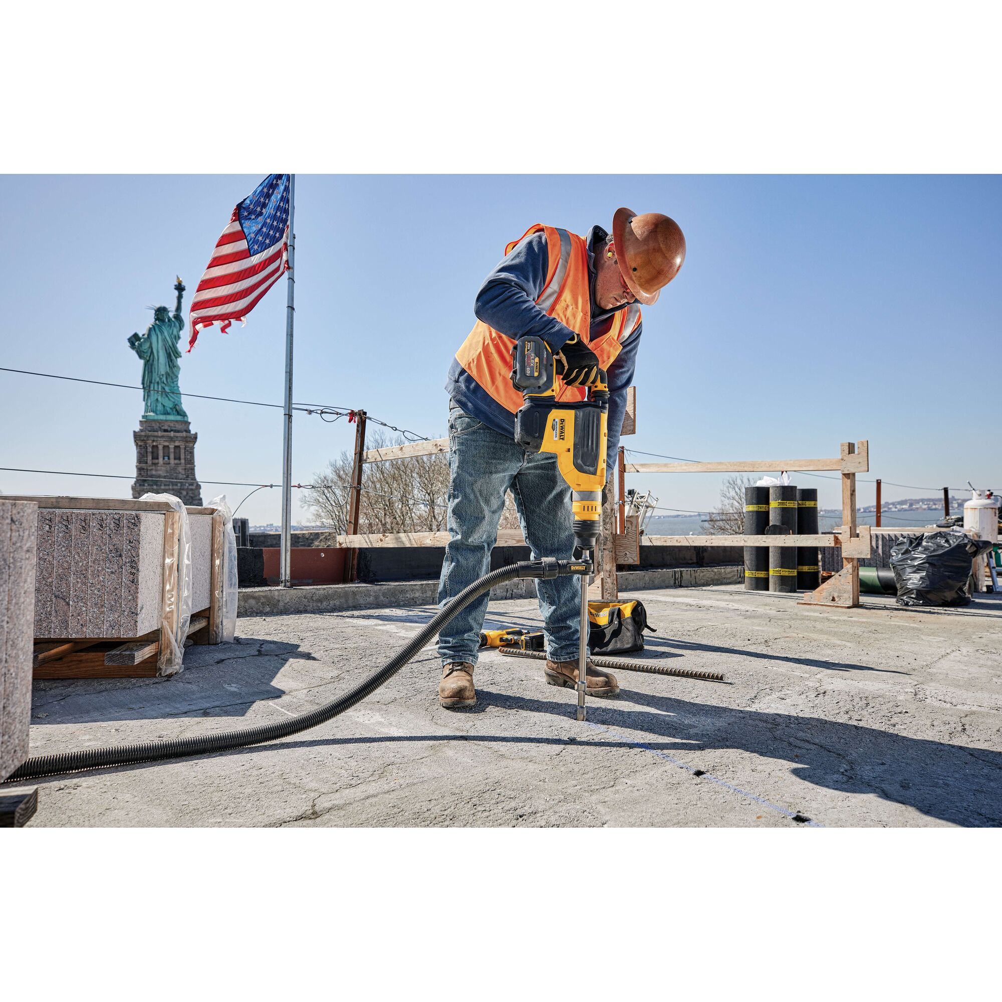 Worker wearing a hard hat and orange safety vest using a yellow DEWALT power tool to drill concrete at a construction site with the Statue of Liberty and an American flag in the background.