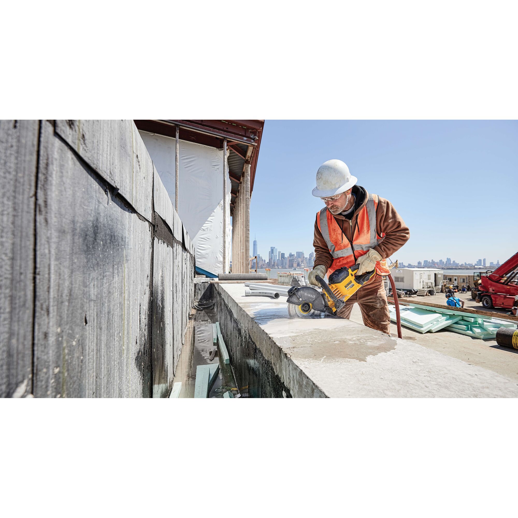 A construction worker wearing a white hard hat and orange safety vest operates a DEWALT power tool to cut concrete on a building site. The city's skyline is visible in the background.