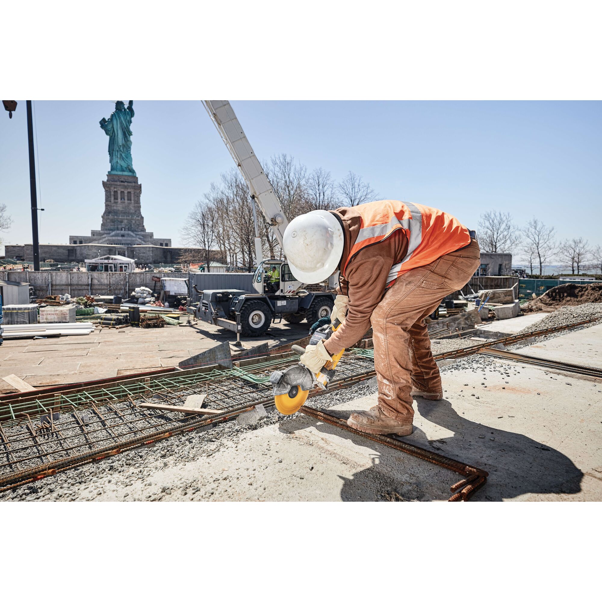 A construction worker wearing a hard hat and orange safety vest uses a DEWALT power tool to cut metal on a construction site, with the Statue of Liberty visible in the background.