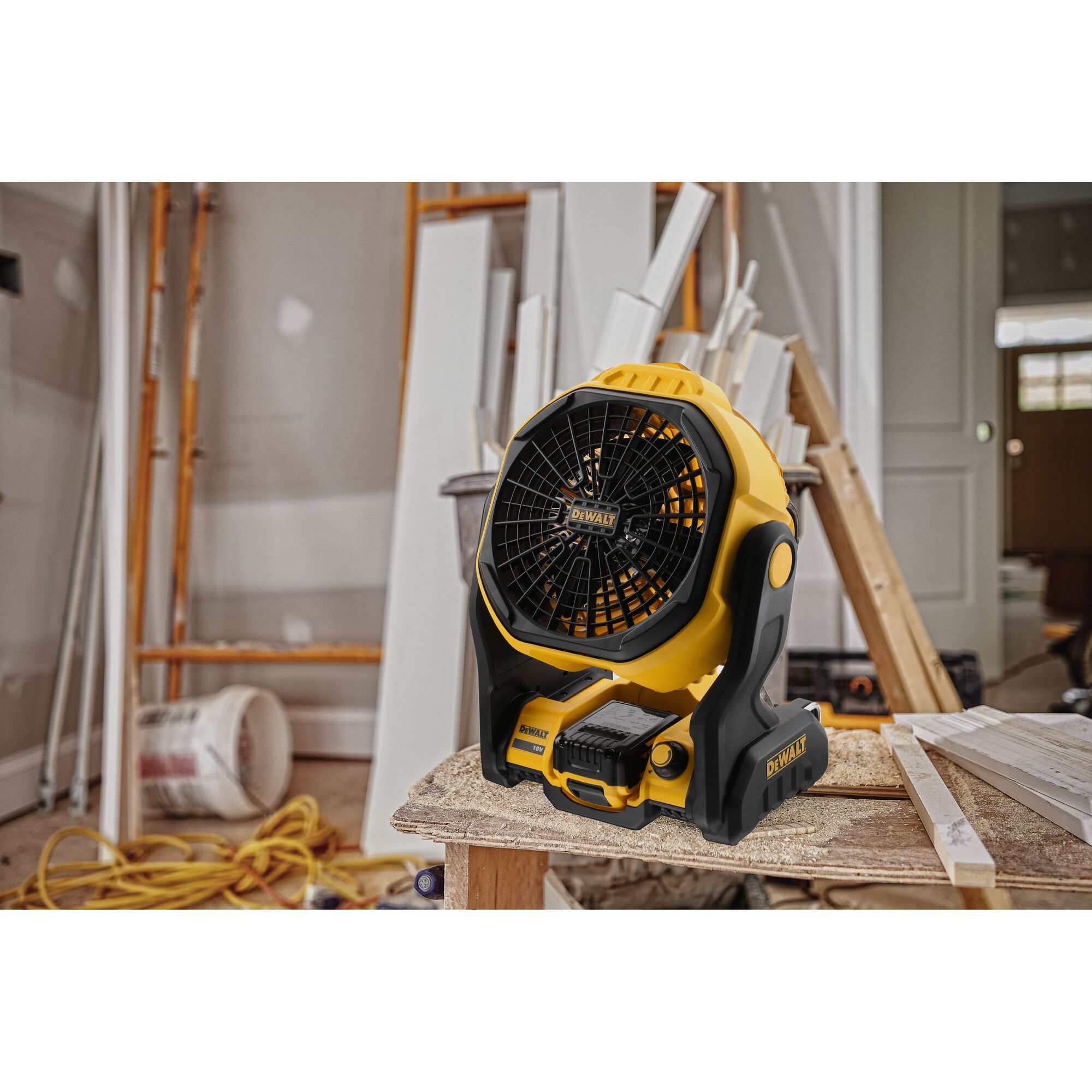 A DEWALT portable battery-powered fan placed on a wooden workbench in a construction or renovation environment, surrounded by tools, wooden planks, and construction materials.
