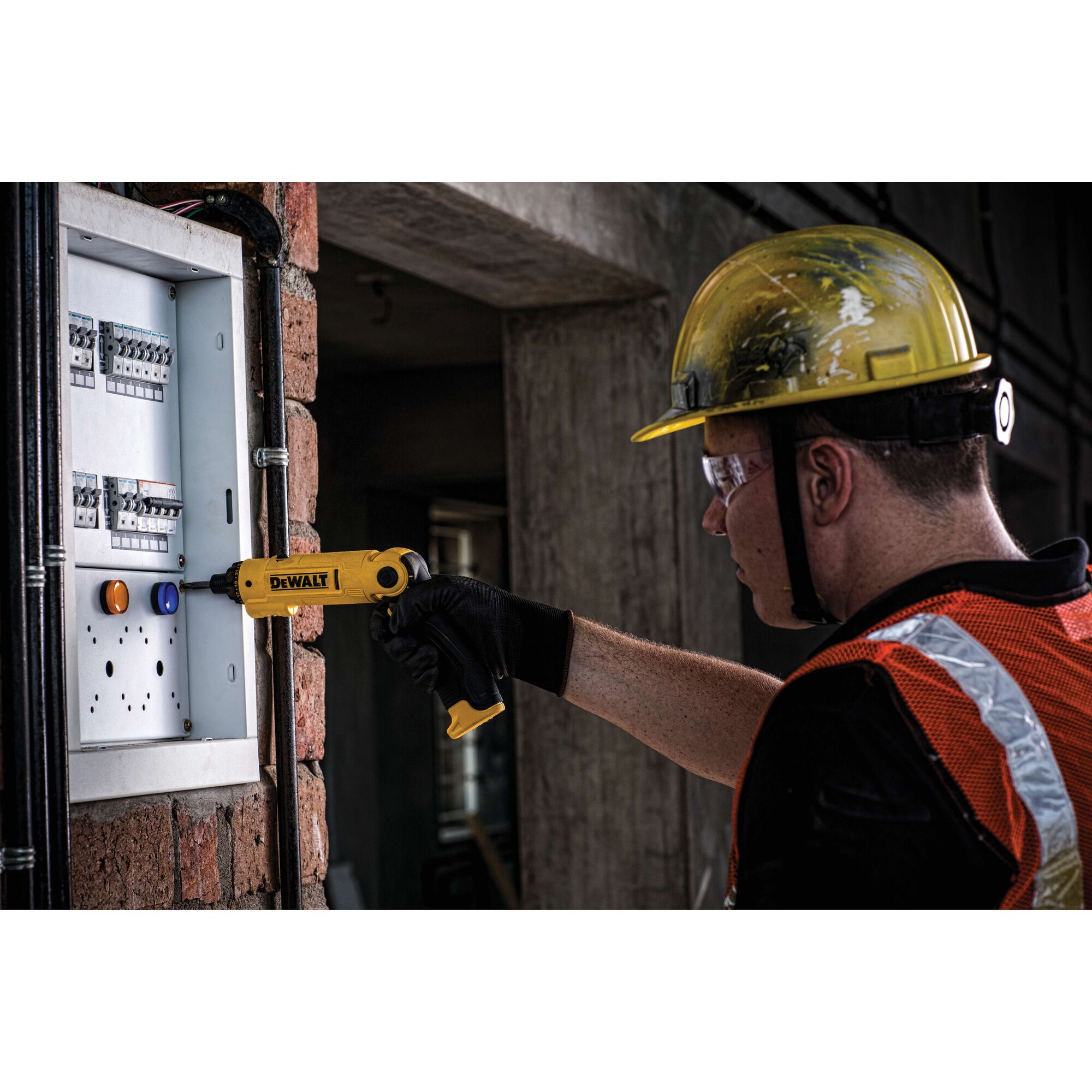 A construction worker wearing a yellow hard hat and orange safety vest uses a yellow DEWALT power tool to work on an electrical panel mounted on a brick wall.