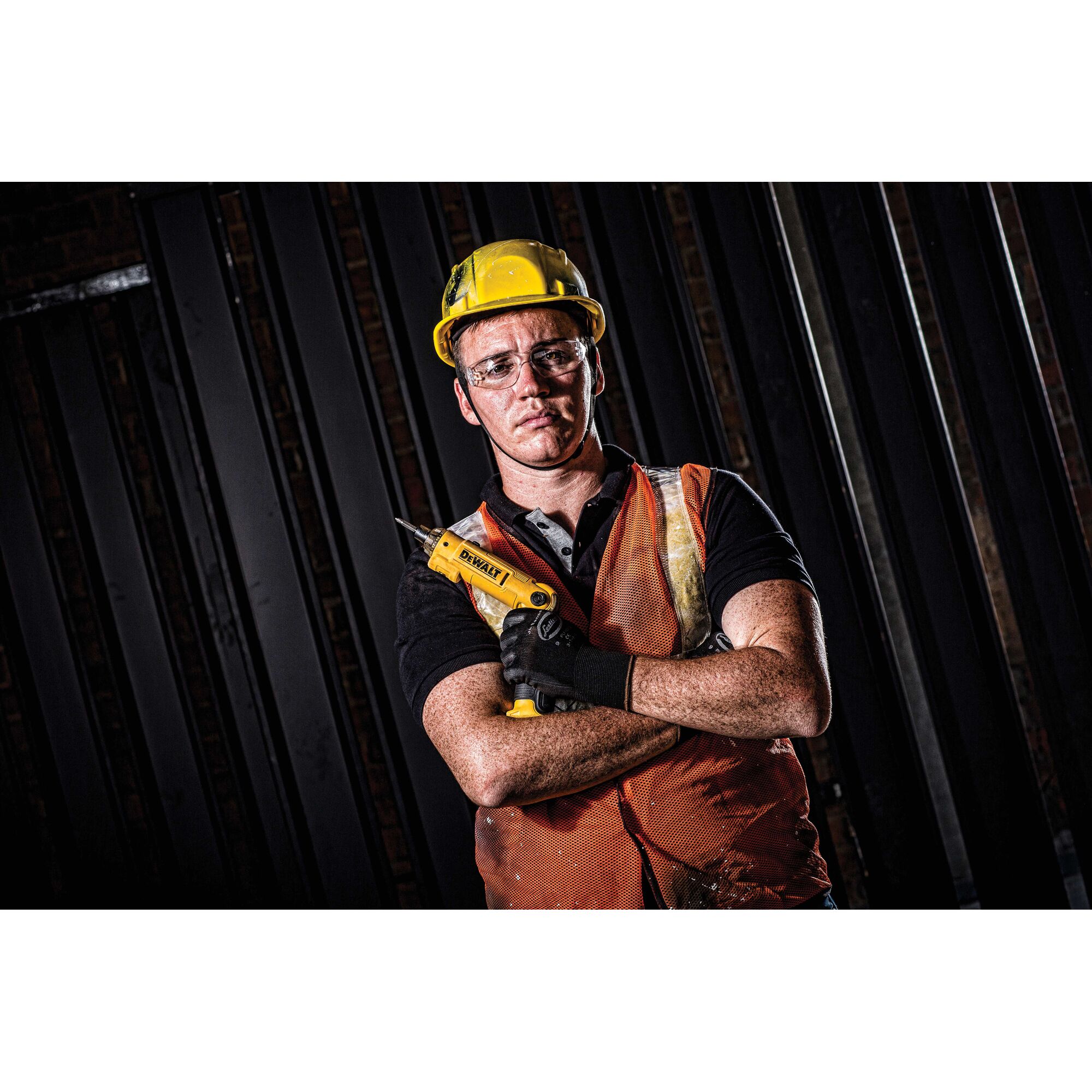 A construction worker wearing a yellow hard hat, orange safety vest, and gloves is holding a DEWALT power tool with arms crossed. The background features vertical black metal beams. The person's face is blurred.