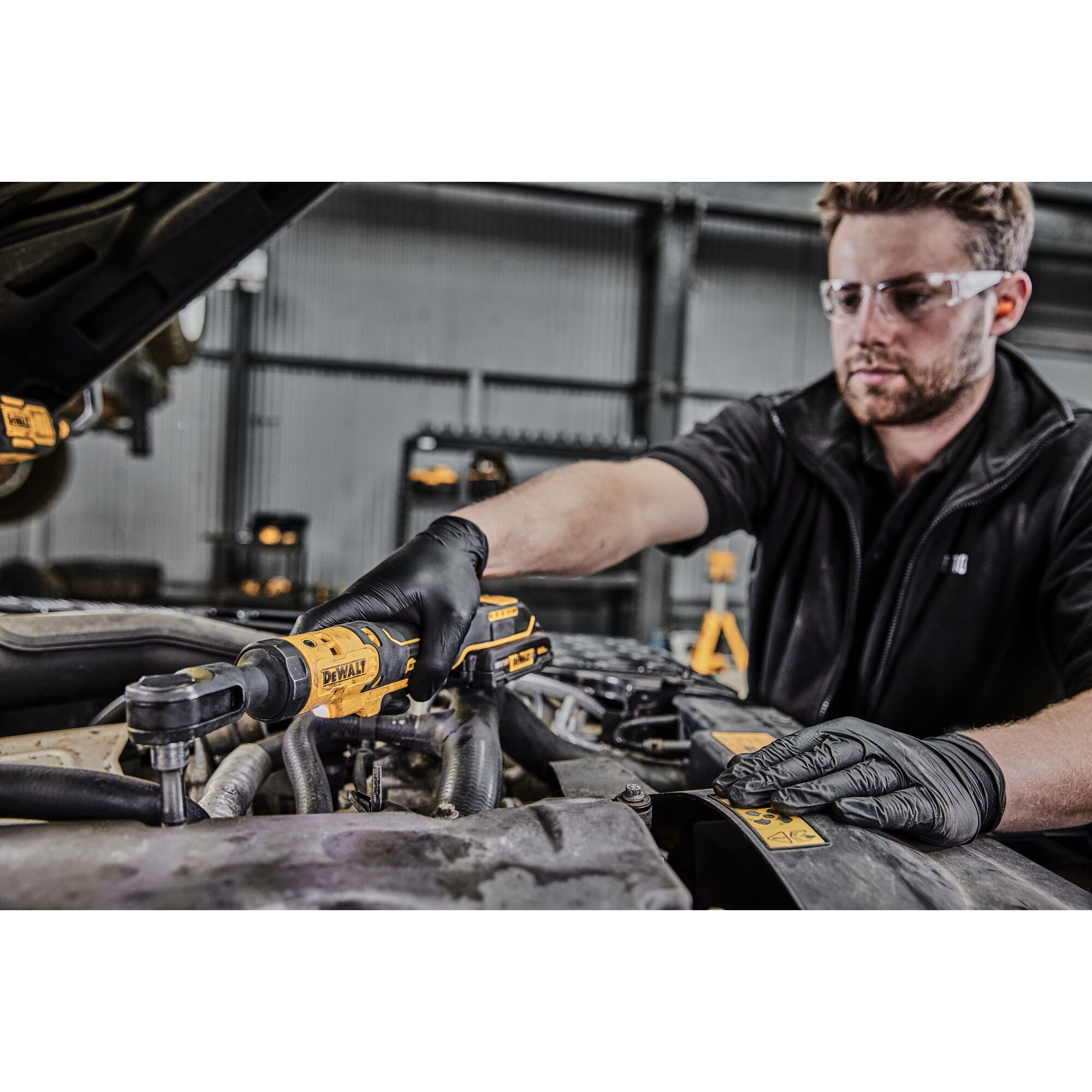 A person wearing black gloves uses a DEWALT DCF512N cordless ratchet tool to work on the engine of a vehicle. Their face is