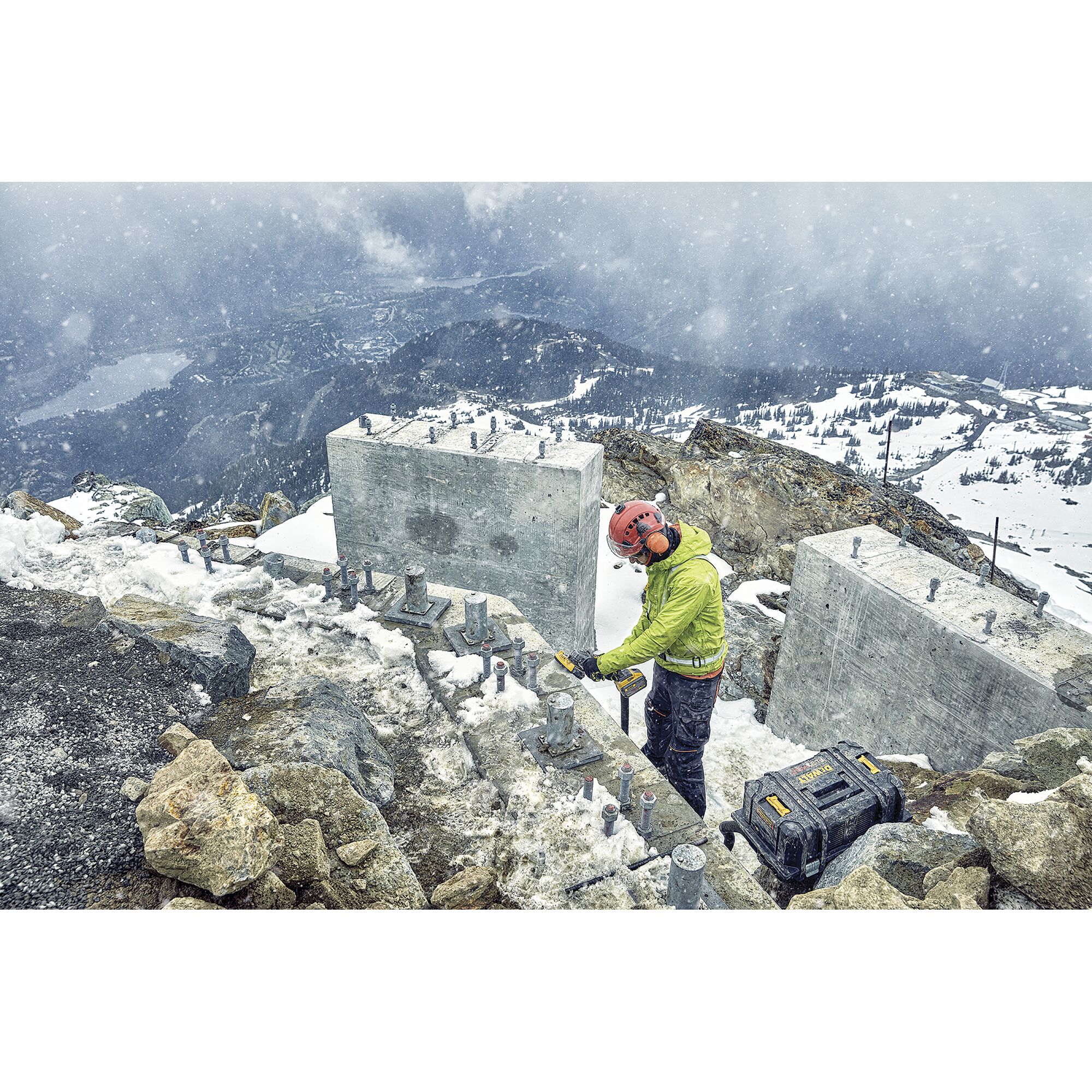 A construction worker in safety gear using a DEWALT tool on a snowy mountain site. Concrete blocks, bolts, and a DEWALT