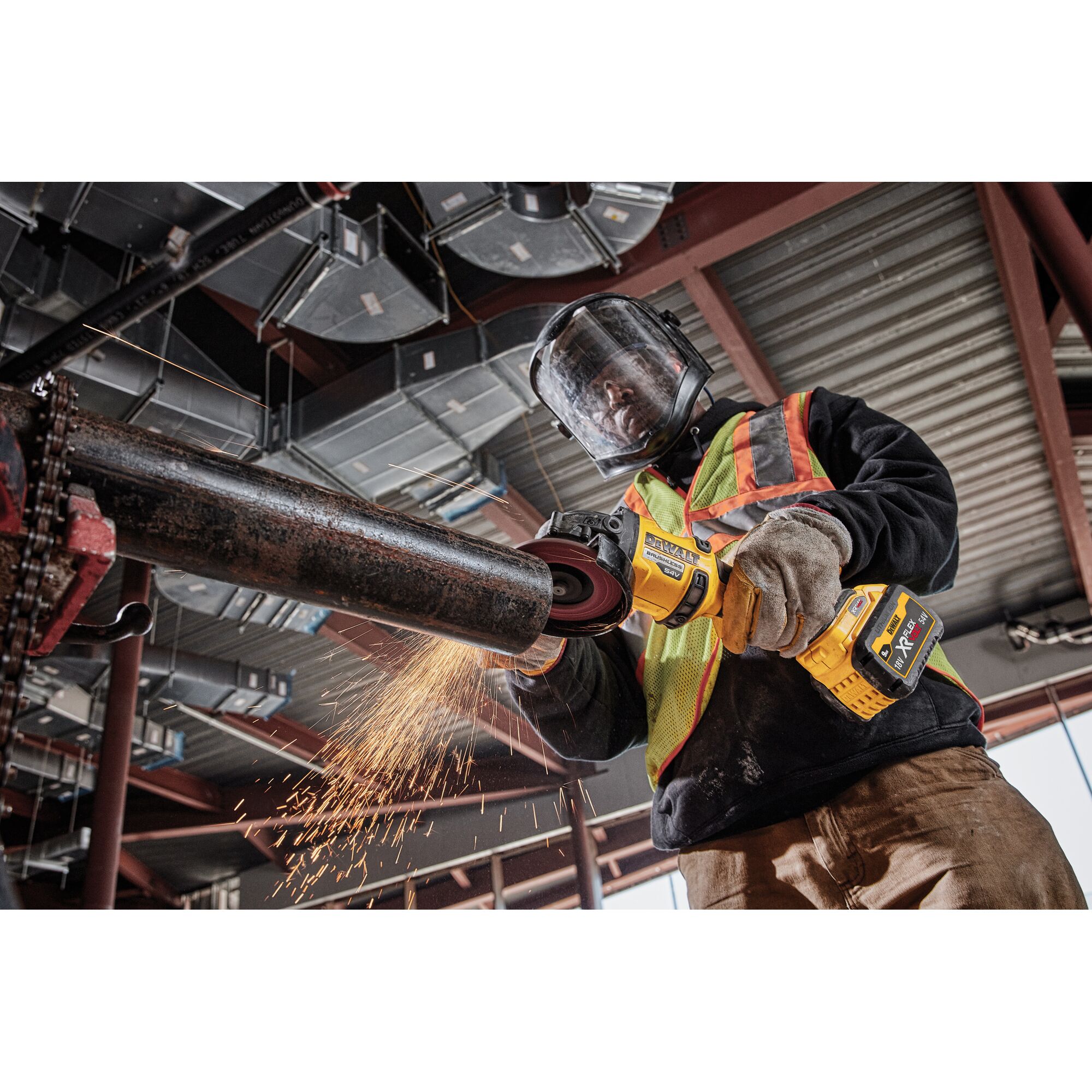 A worker wearing safety gear uses a DEWALT DCG418NT cordless angle grinder to cut a metal pipe, producing sparks in an