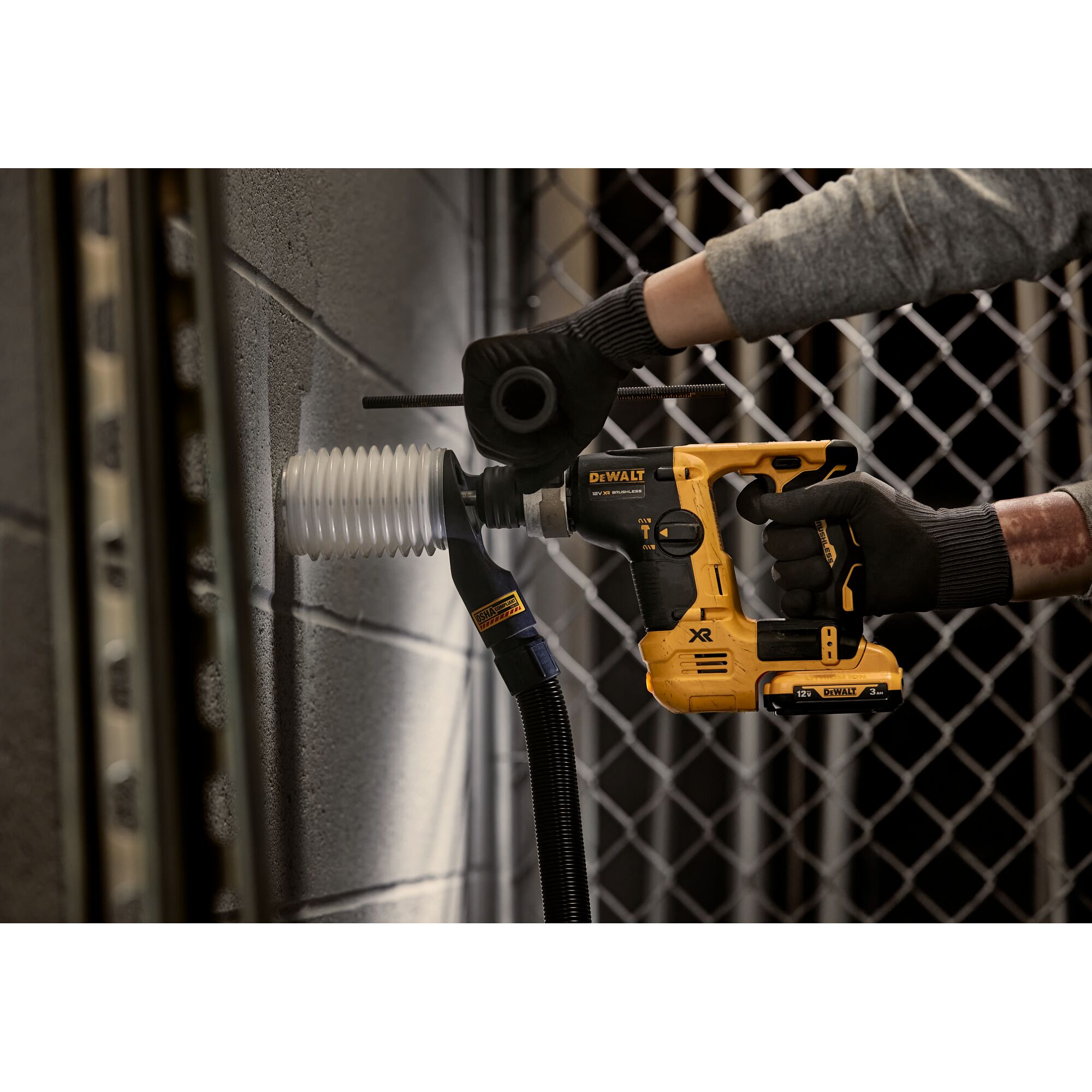 A person wearing gloves uses a DEWALT cordless rotary hammer drill (model DCH072L2) with a dust extraction attachment to drill into a concrete wall.