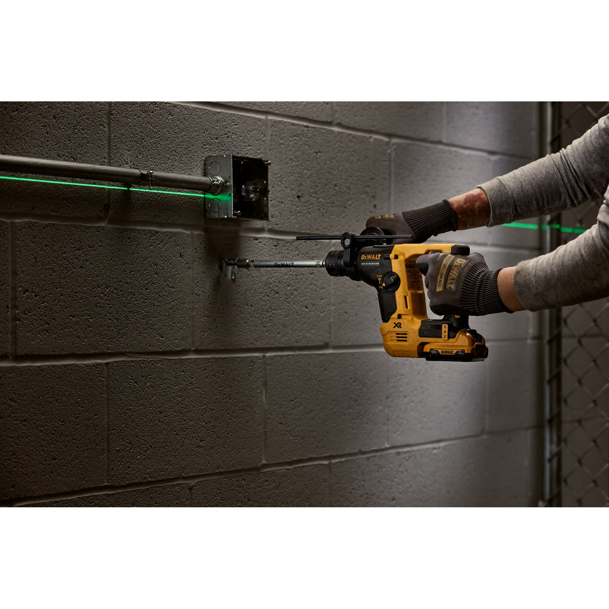 A person wearing gloves uses a DEWALT cordless rotary hammer drill to install or drill into a concrete block wall. The drill is yellow and black, and there is a green laser line visible across metal conduit for alignment.