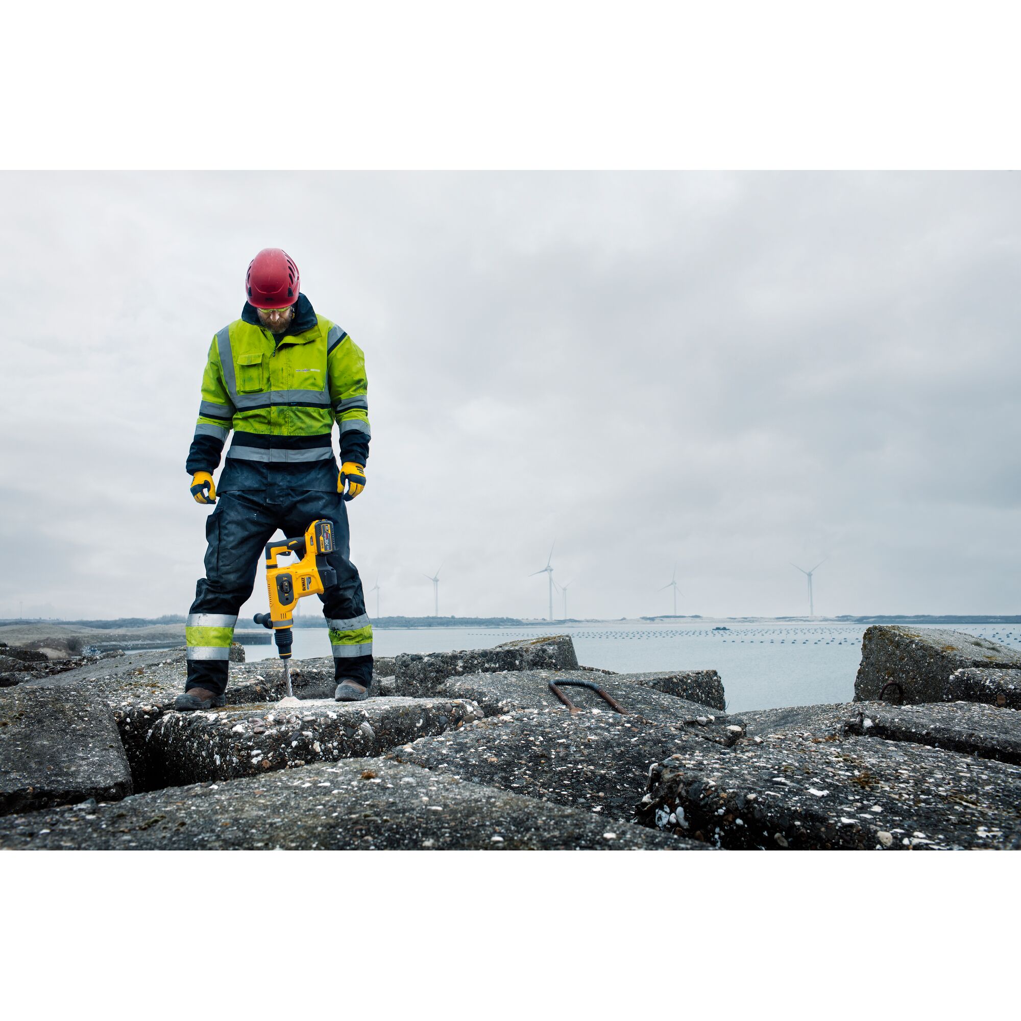 A person wearing high-visibility workwear and a red helmet is using a yellow DEWALT rotary hammer drill (SKU: DCH481N) to drill into concrete at an outdoor construction site by the water, with wind turbines visible in the background.