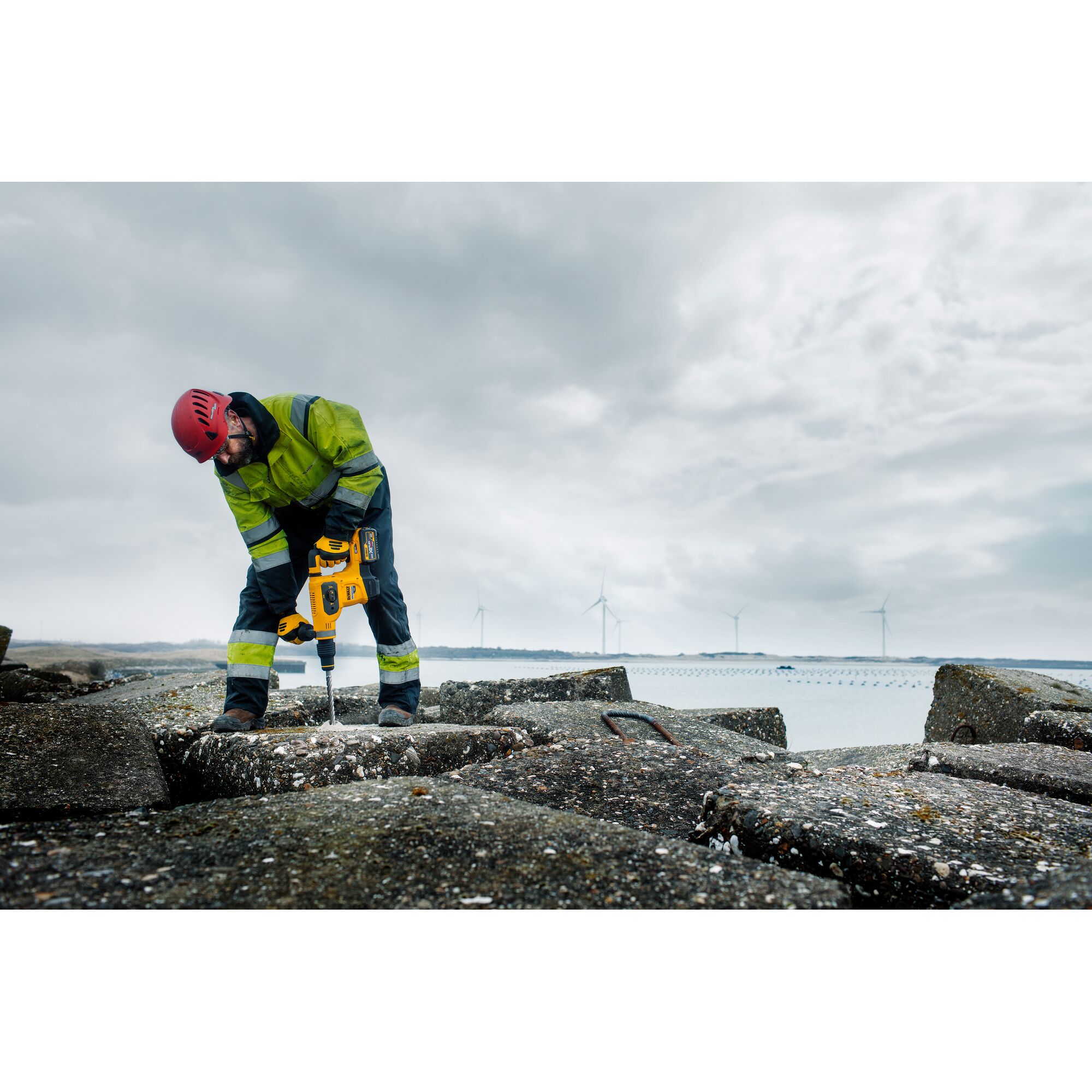 Worker using yellow DEWALT tool outdoors on concrete, wearing safety gear. Wind turbines in background.