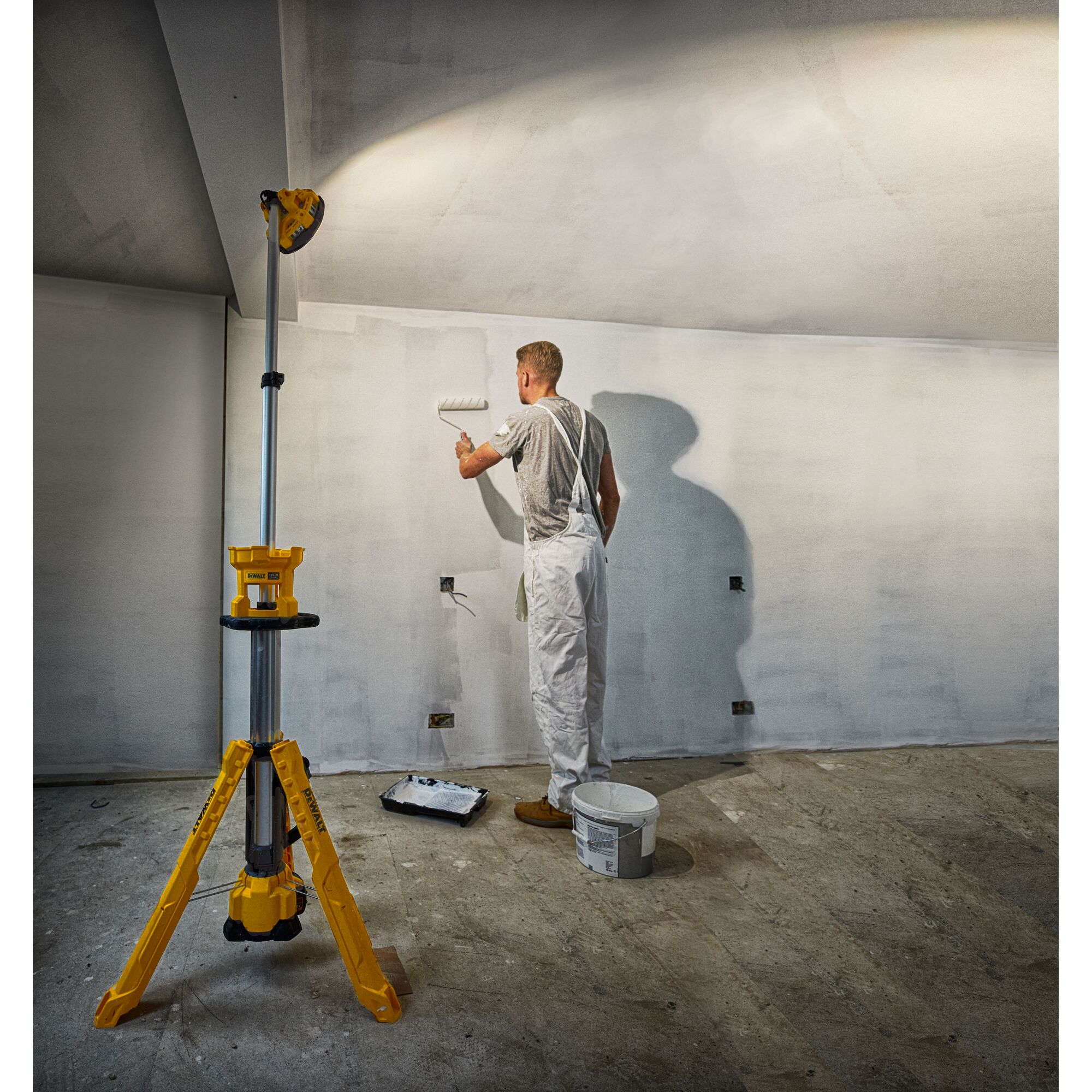 A painter uses a paint roller on a wall, illuminated by a yellow DEWALT tripod work light in a room under renovation.