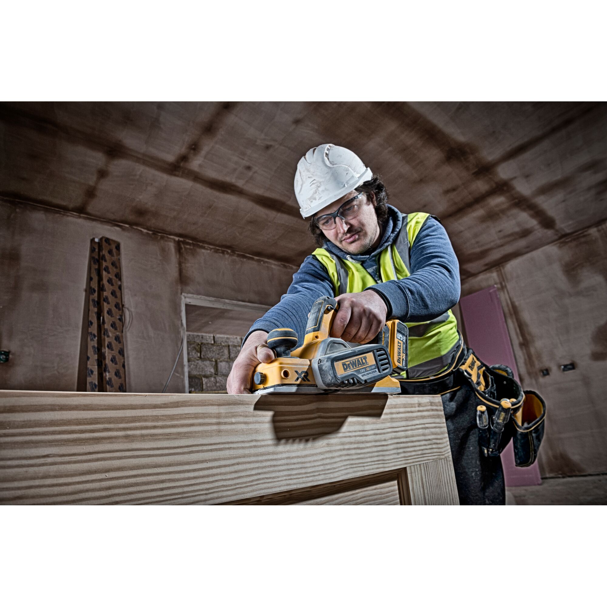 A construction worker wearing a hard hat and safety vest is using a DEWALT DCP580N cordless planer on a wooden surface inside a building under construction.