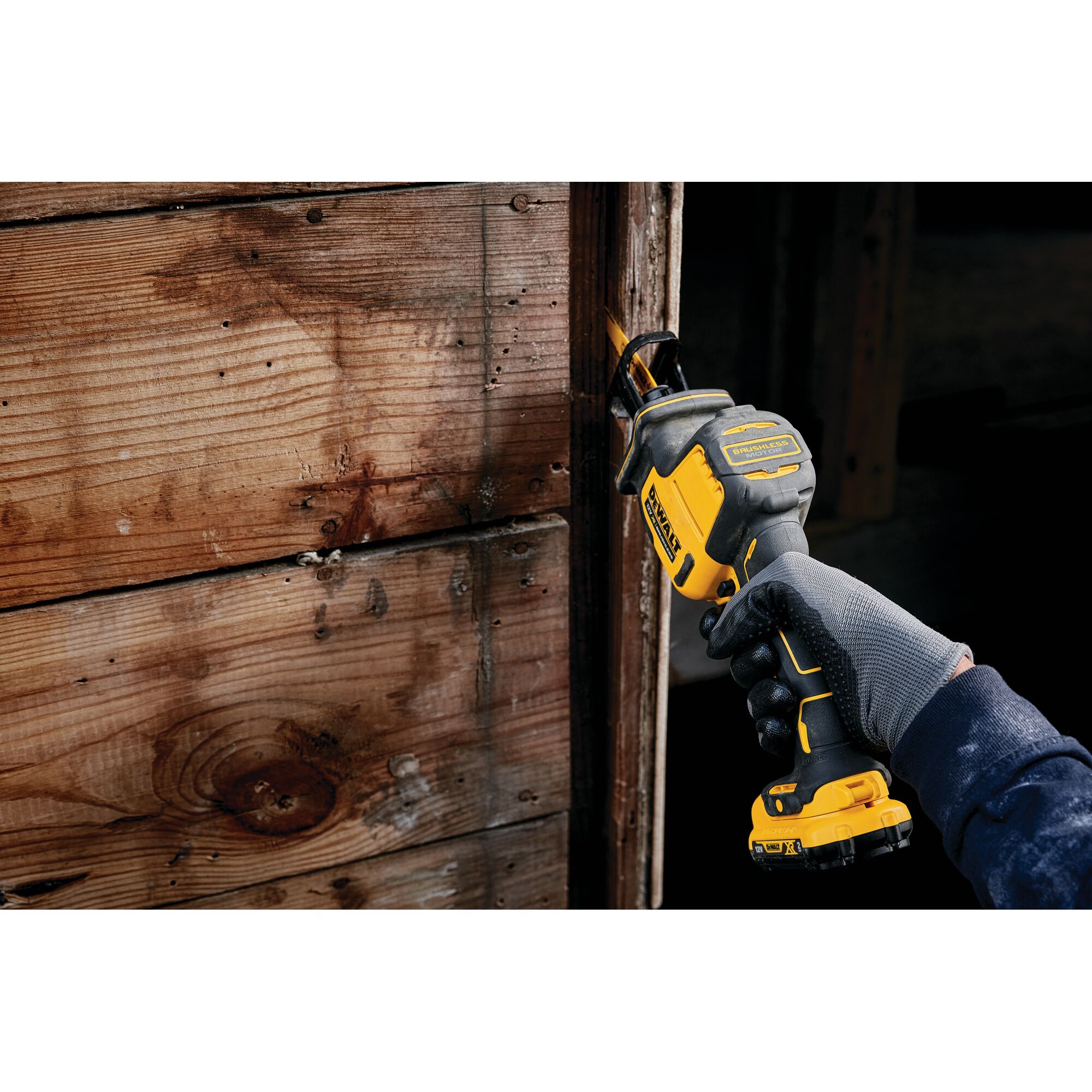 Gloved hand using a DEWALT cordless reciprocating saw on a wooden surface indoors.
