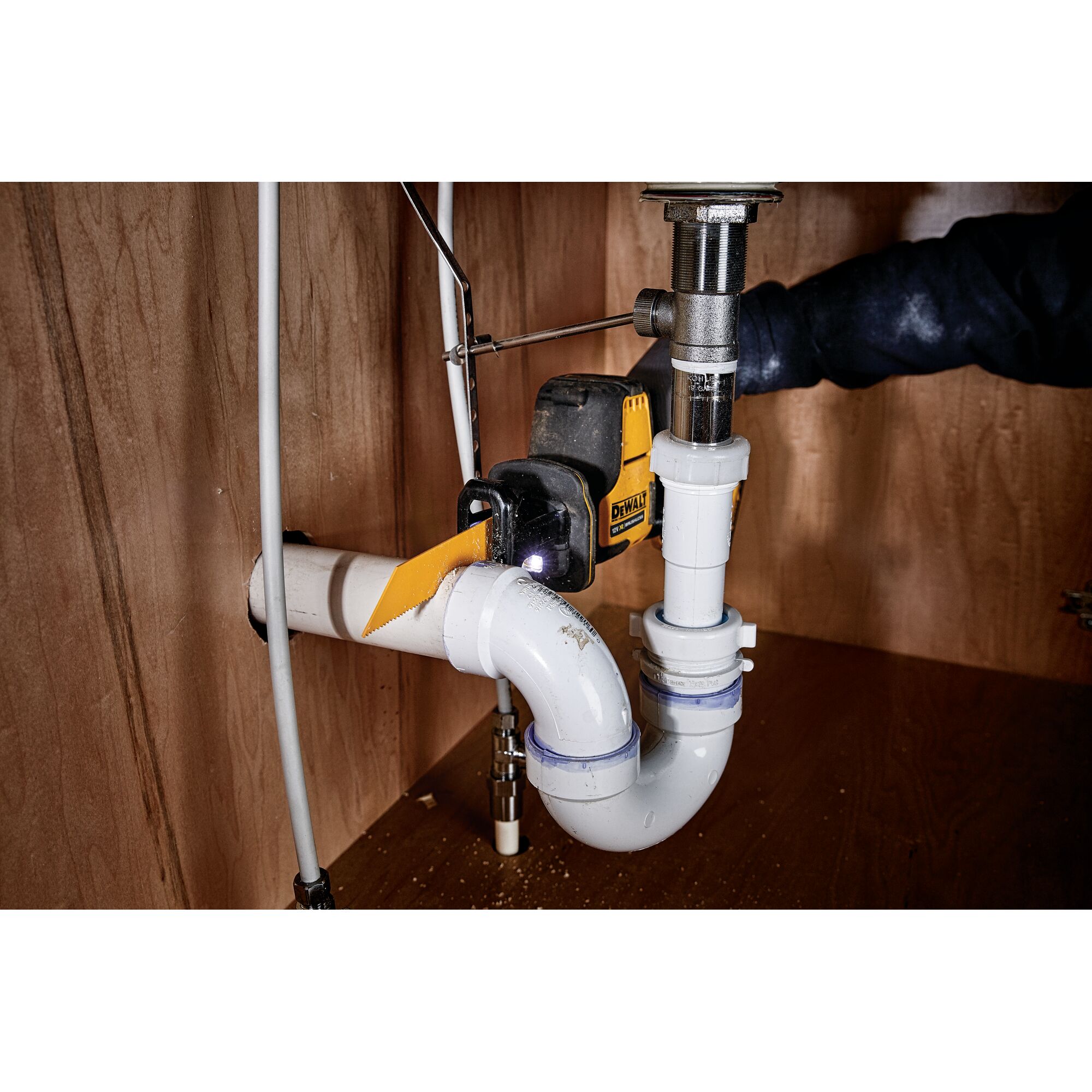 DEWALT cordless saw cutting a PVC pipe under a sink in a wooden cabinet.