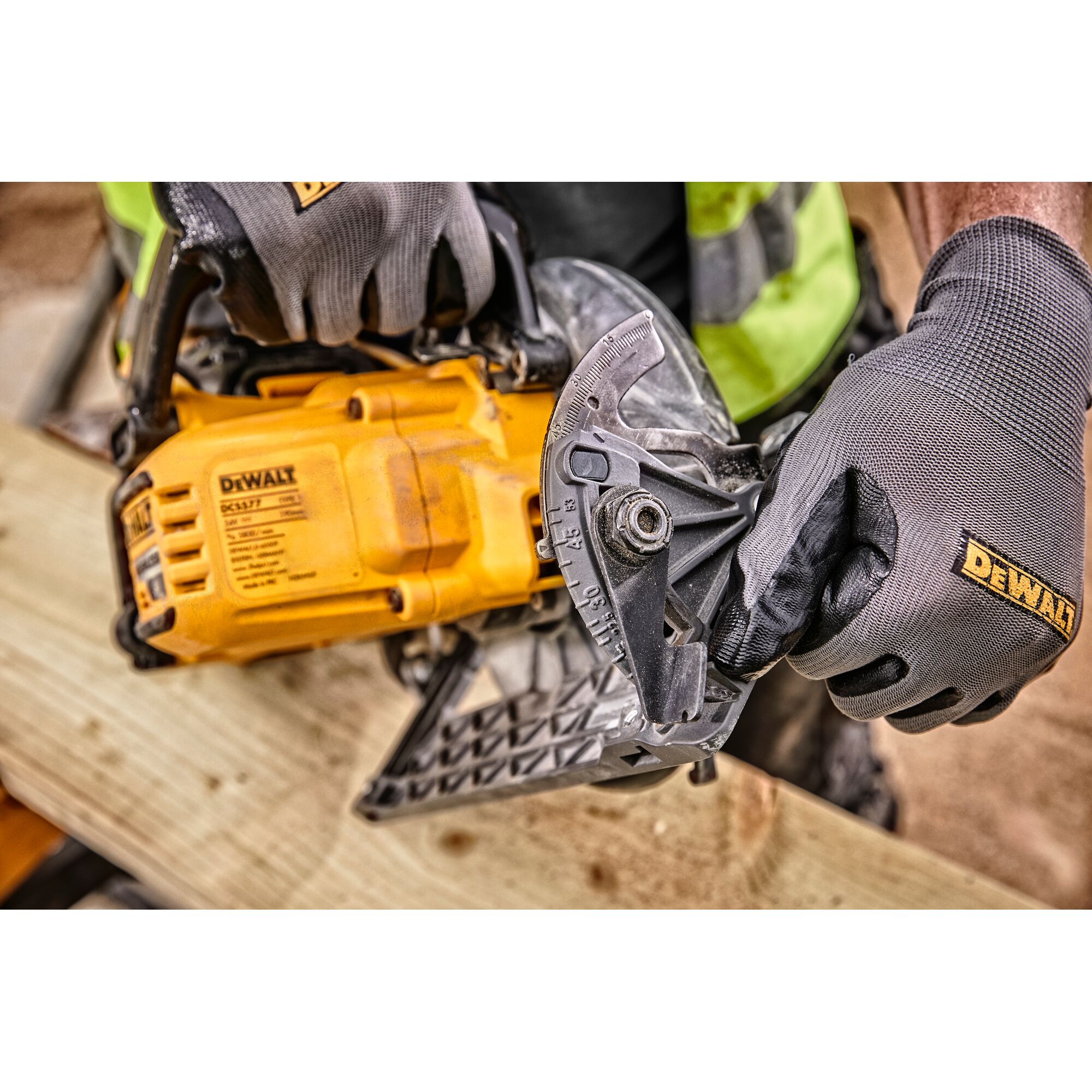 Gloved hands adjust a yellow DEWALT DCS577N saw placed on a wooden board at a construction site.