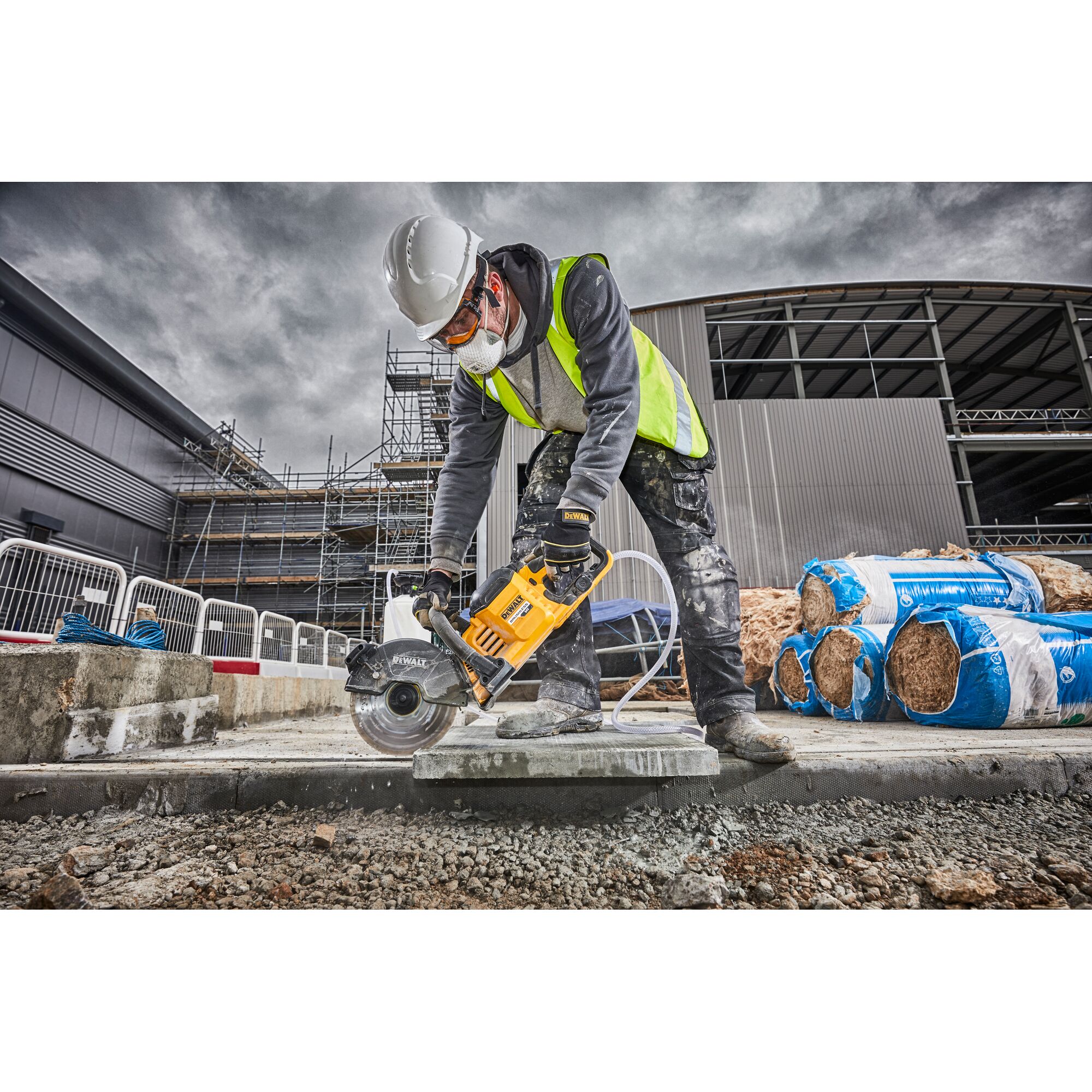 Person using a DEWALT power saw to cut concrete at a construction site, wearing safety gear and a high-visibility vest.