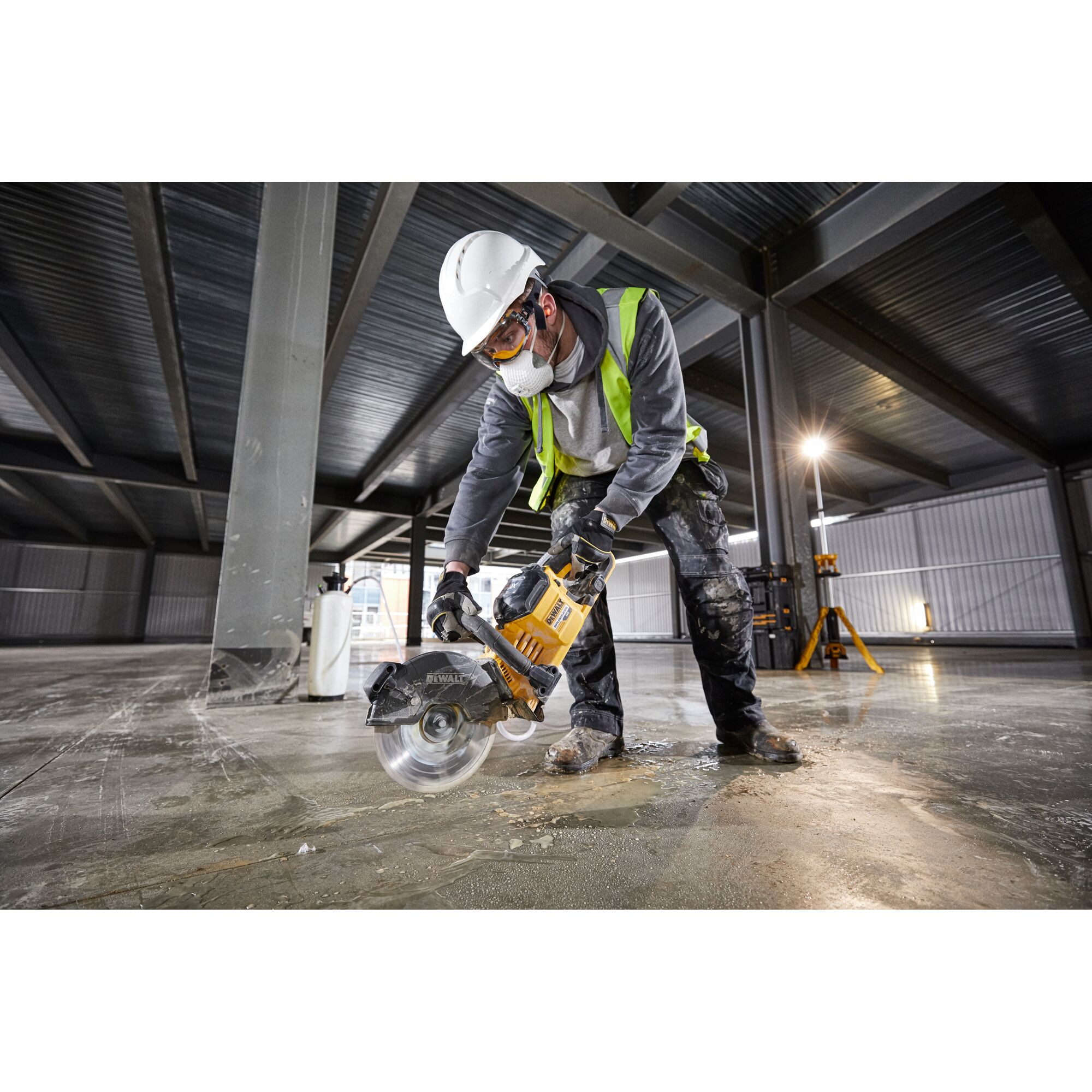 Worker using a DEWALT DCS690N saw to cut concrete on a construction site, wearing safety gear and high-visibility vest.
