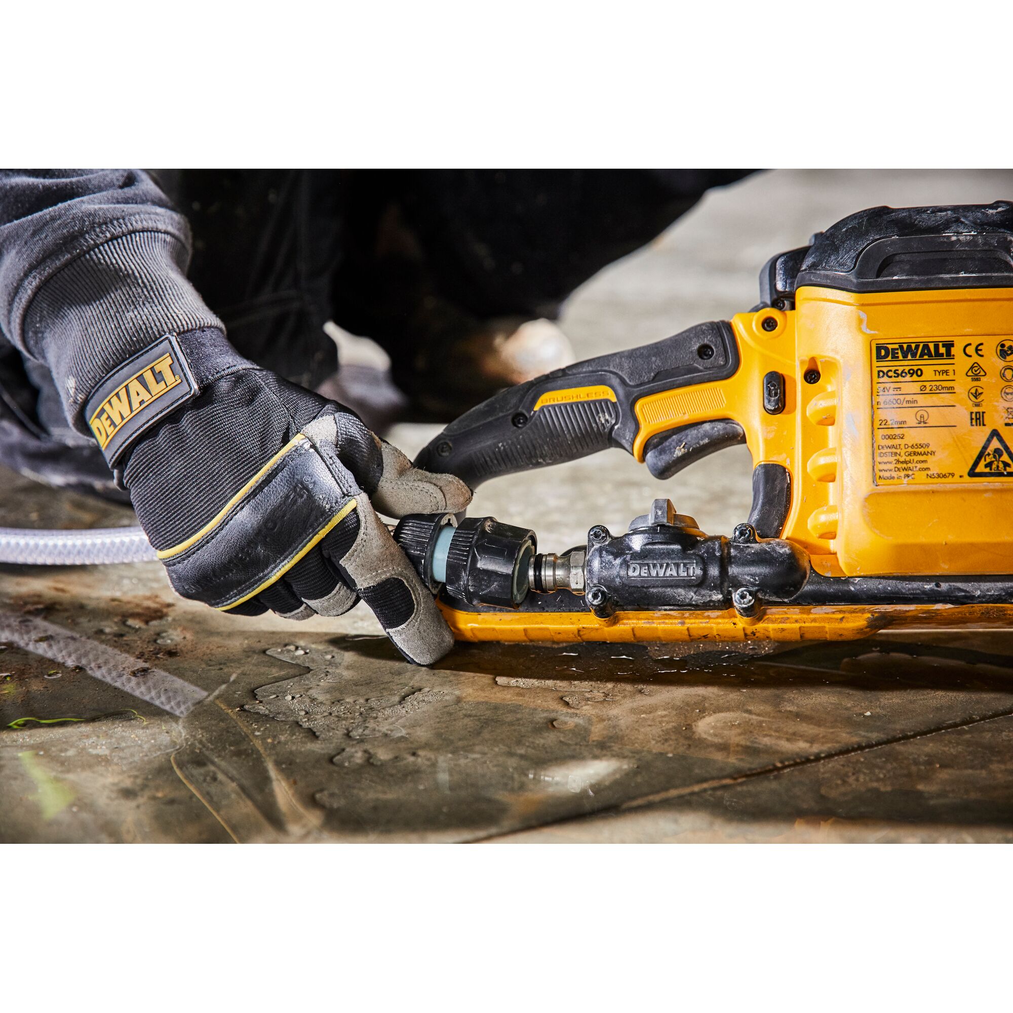 Gloved hand connects hose to a yellow DEWALT DCS690 power tool placed on a wet concrete surface.