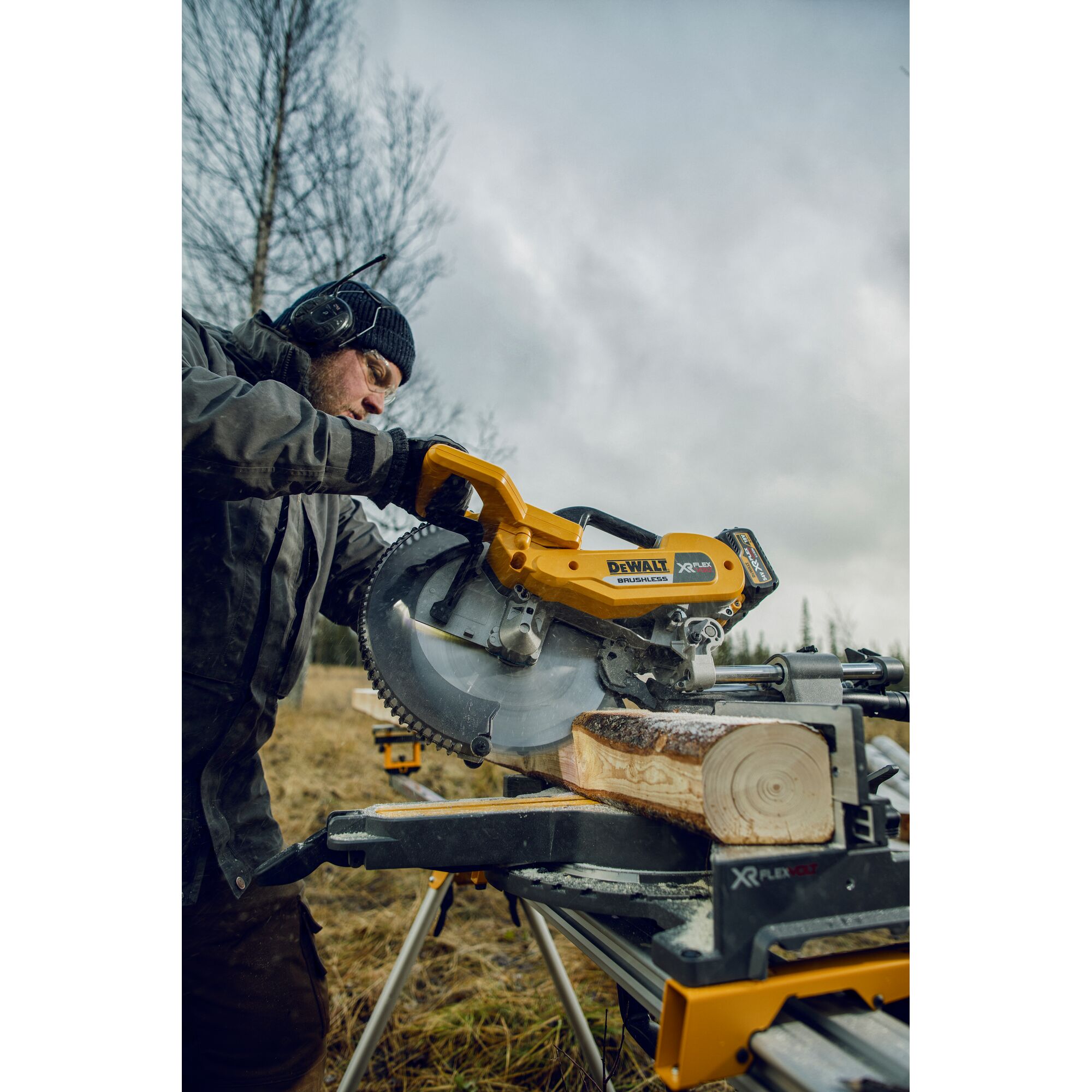 A person using a DEWALT DHS780T2 cordless miter saw outdoors to cut a piece of wood. The person is wearing safety gear, including hearing protection, and the tool is positioned on a stand in a grassy area with trees in the background.
