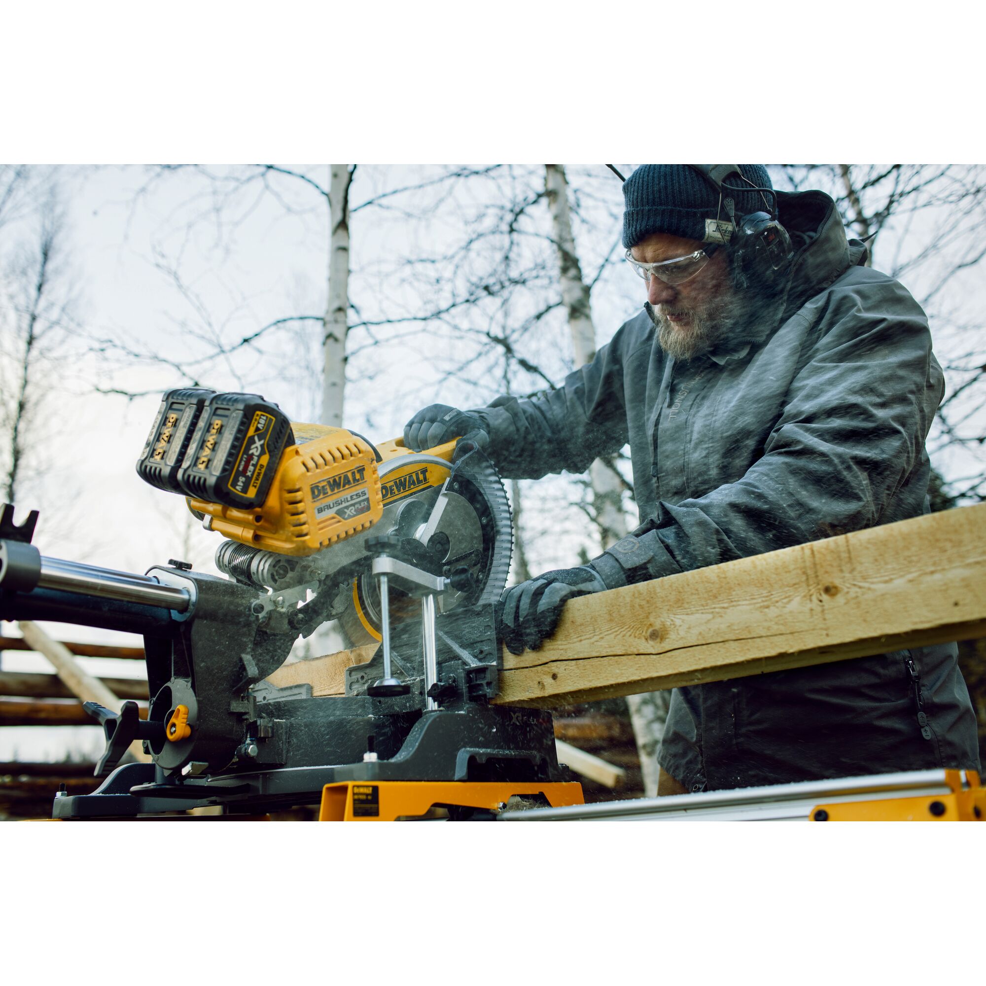 A person wearing protective clothing is using a DEWALT DHS780T2 cordless sliding miter saw to cut a wooden board outdoors. The saw is yellow and black with visible dual battery packs, and wood dust is being generated during the cut.
