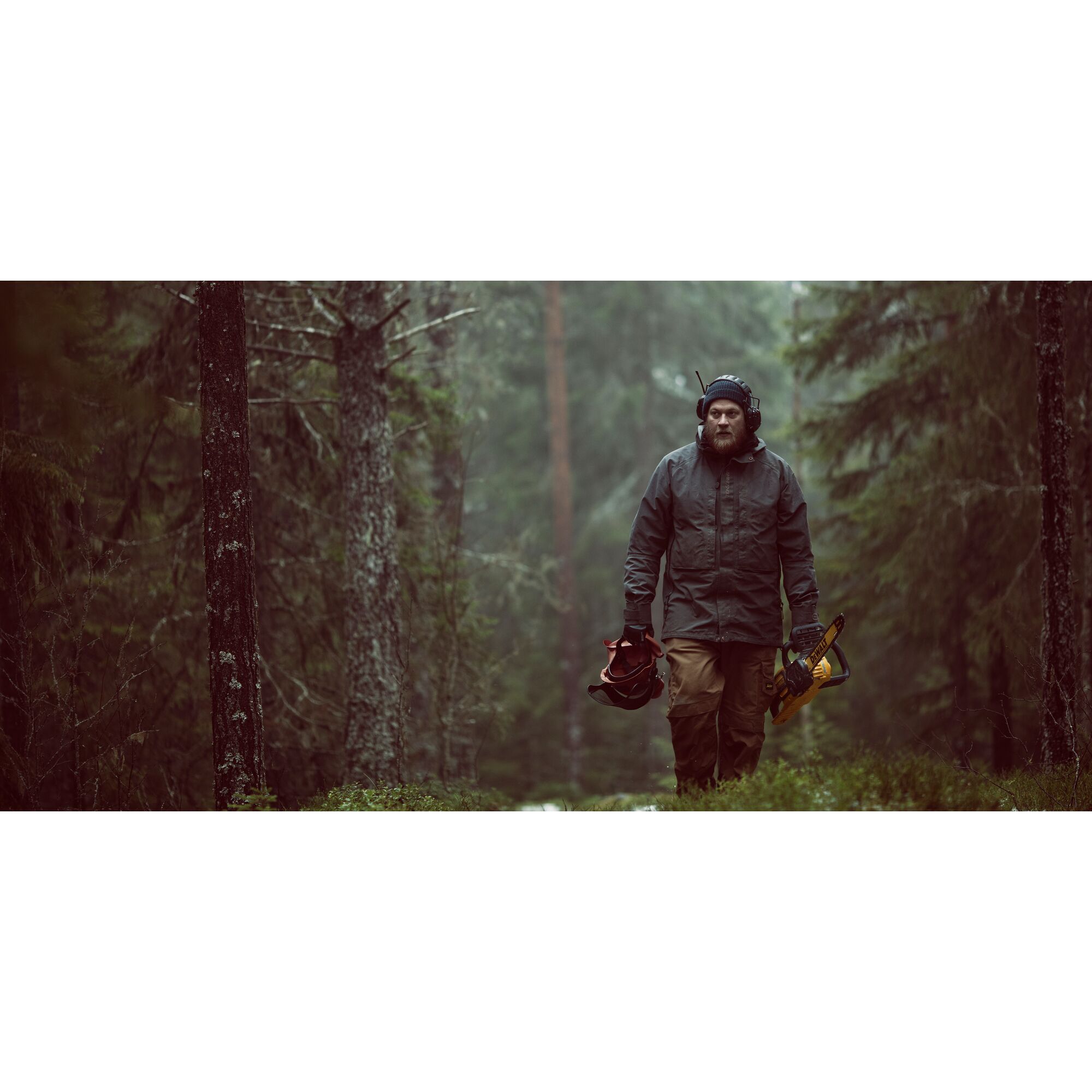 A person wearing outdoor work clothing and protective headgear walks through a forest, holding a DEWALT chainsaw in one hand and additional equipment in the other. The scene is misty and surrounded by tall trees.