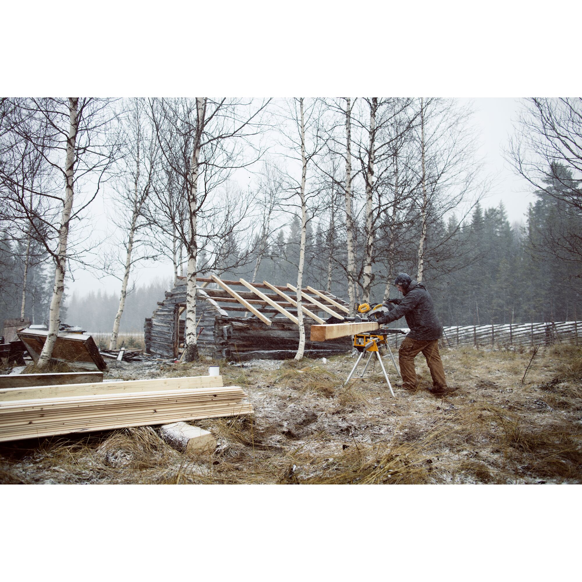 A person using a DEWALT miter saw outdoors in a snowy, wooded area, cutting a large piece of wood. There is a partially constructed wooden structure and stacks of lumber nearby.