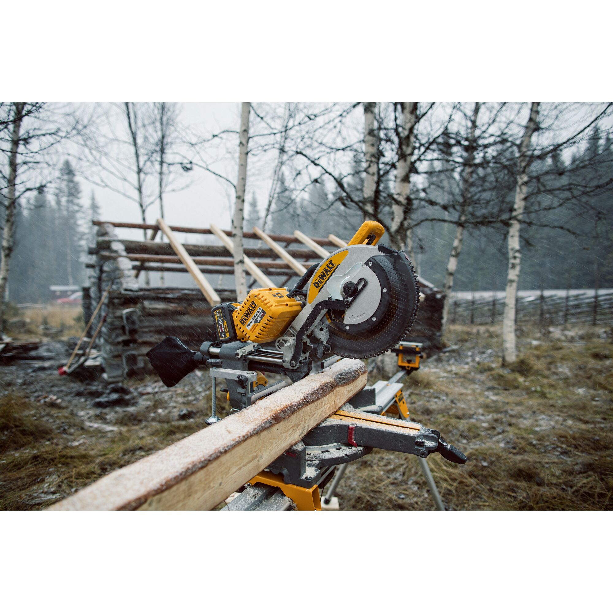 A DEWALT DHS780T2 miter saw is set up outdoors on a stand, cutting a frost-covered wooden board. The scene is in a snowy, wooded area with bare trees and a partially built wooden structure in the background.