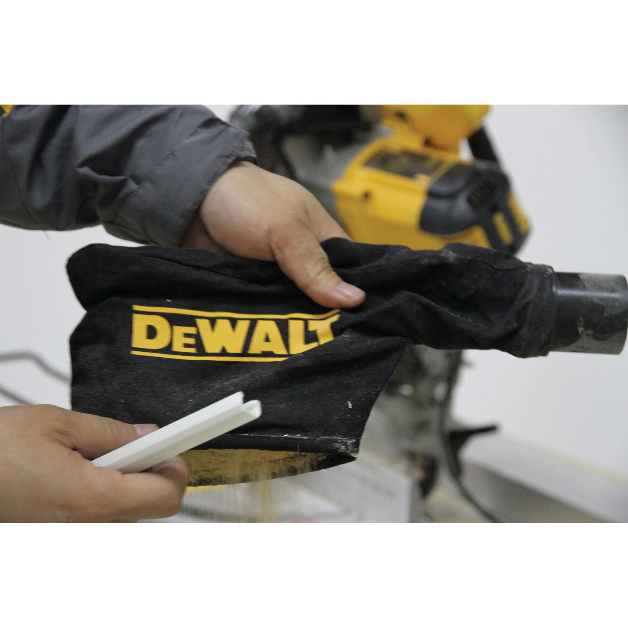 Close-up of a person's hands holding a DEWALT branded dust collection bag attached to a power tool, with part of the yellow tool visible in the background.