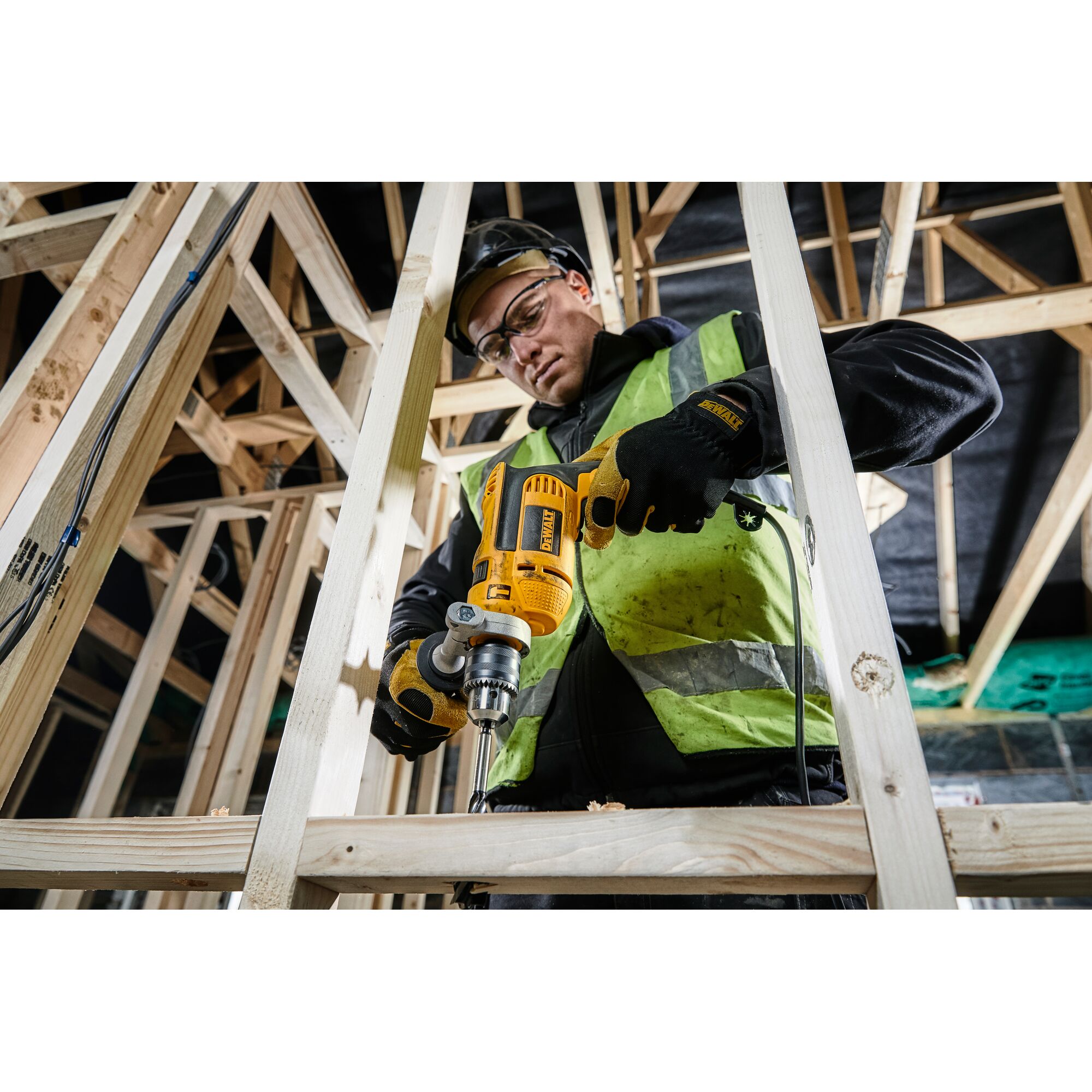 A person wearing a high-visibility vest and gloves uses a yellow DEWALT drill to work on a wooden frame at a construction site.