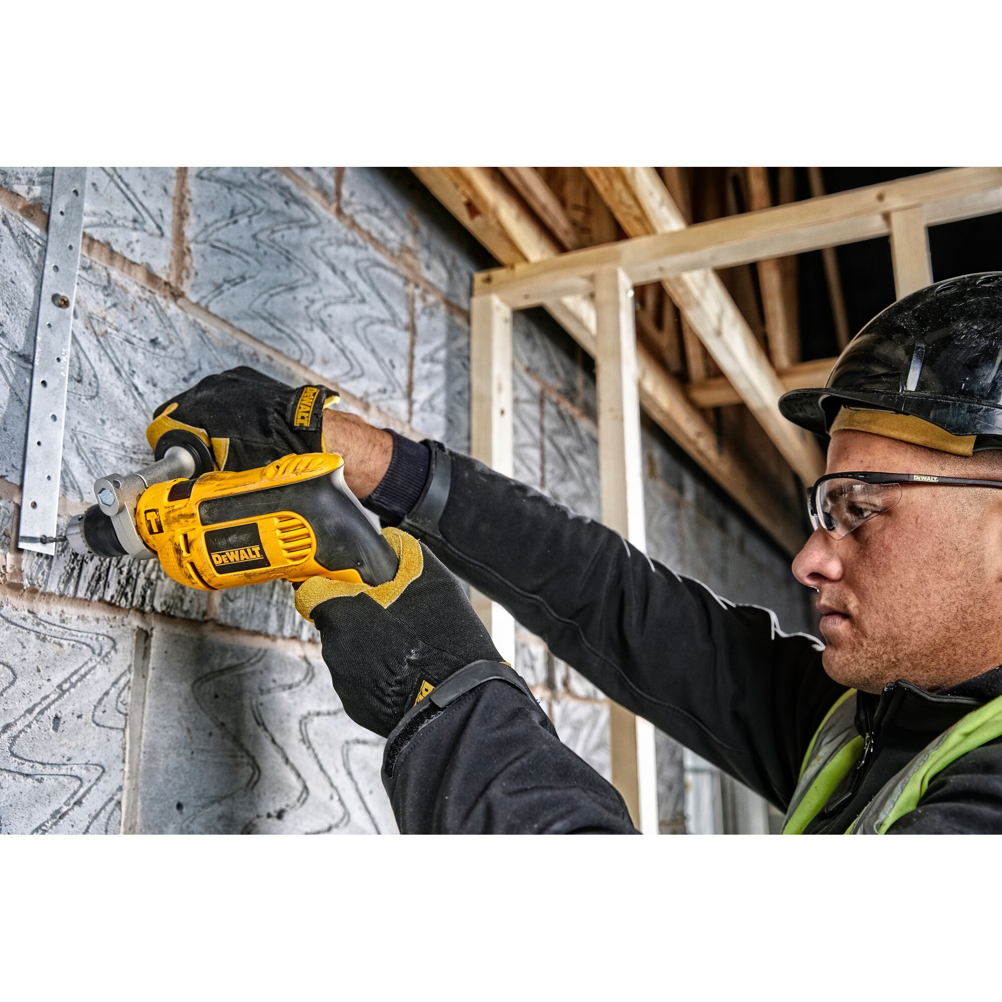 A person wearing protective gloves and a helmet uses a yellow and black DEWALT power drill (model DWD024K) to work on a metal bracket attached to a brick wall at a construction site.