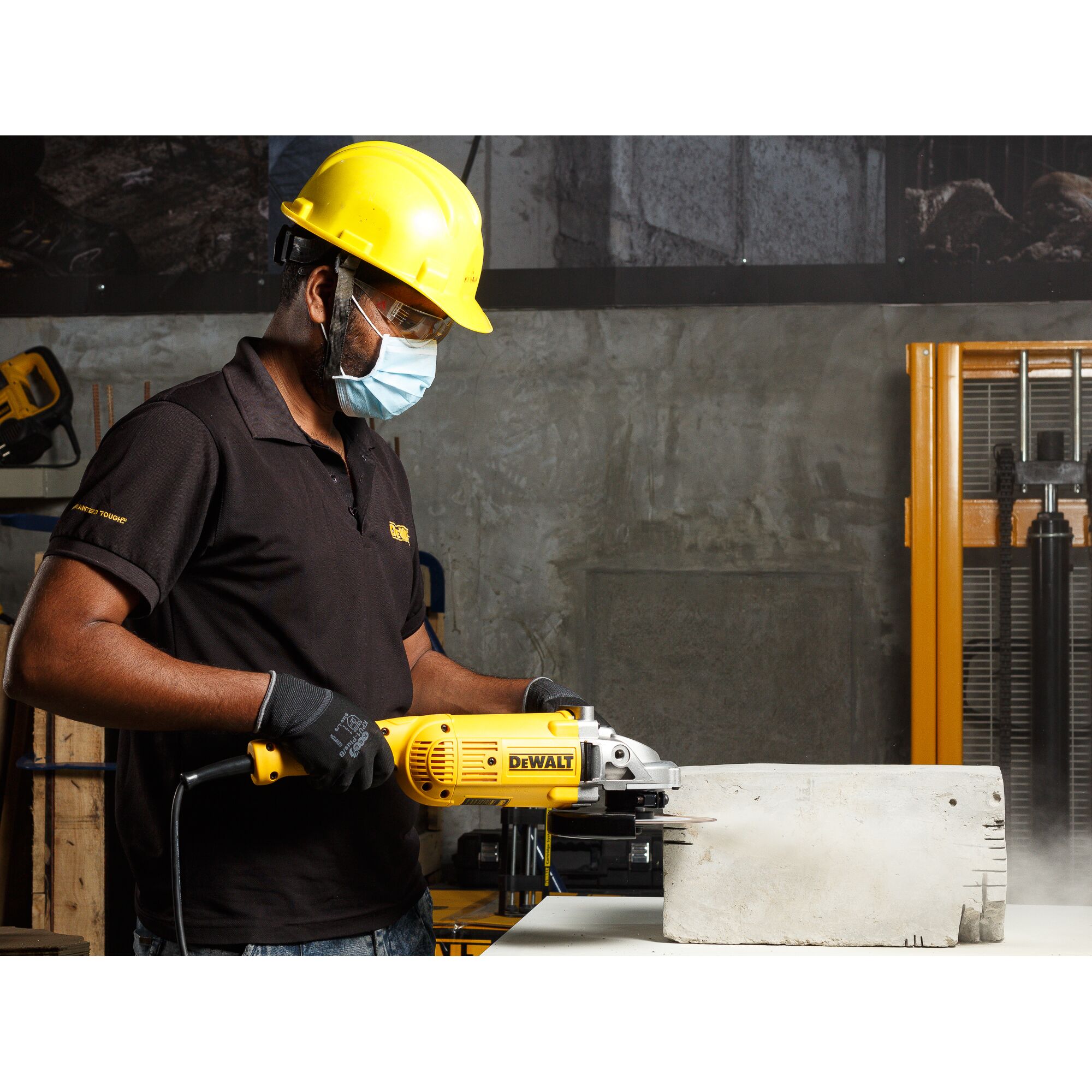 A person wearing a yellow hard hat, black gloves, and a black polo shirt is using a DEWALT DWE493 angle grinder to cut a large concrete block in an industrial workshop setting.