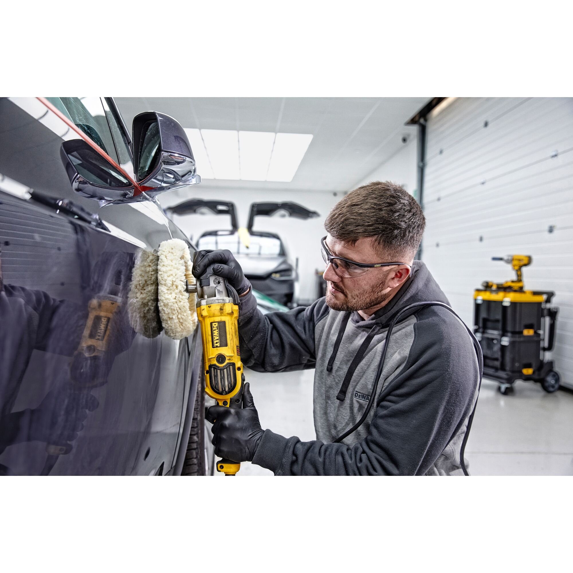 A person wearing gloves uses a DEWALT DWP849X polisher to buff the side of a dark-colored car in a garage. The person's face is blurred, and there are tools and equipment visible in the background.