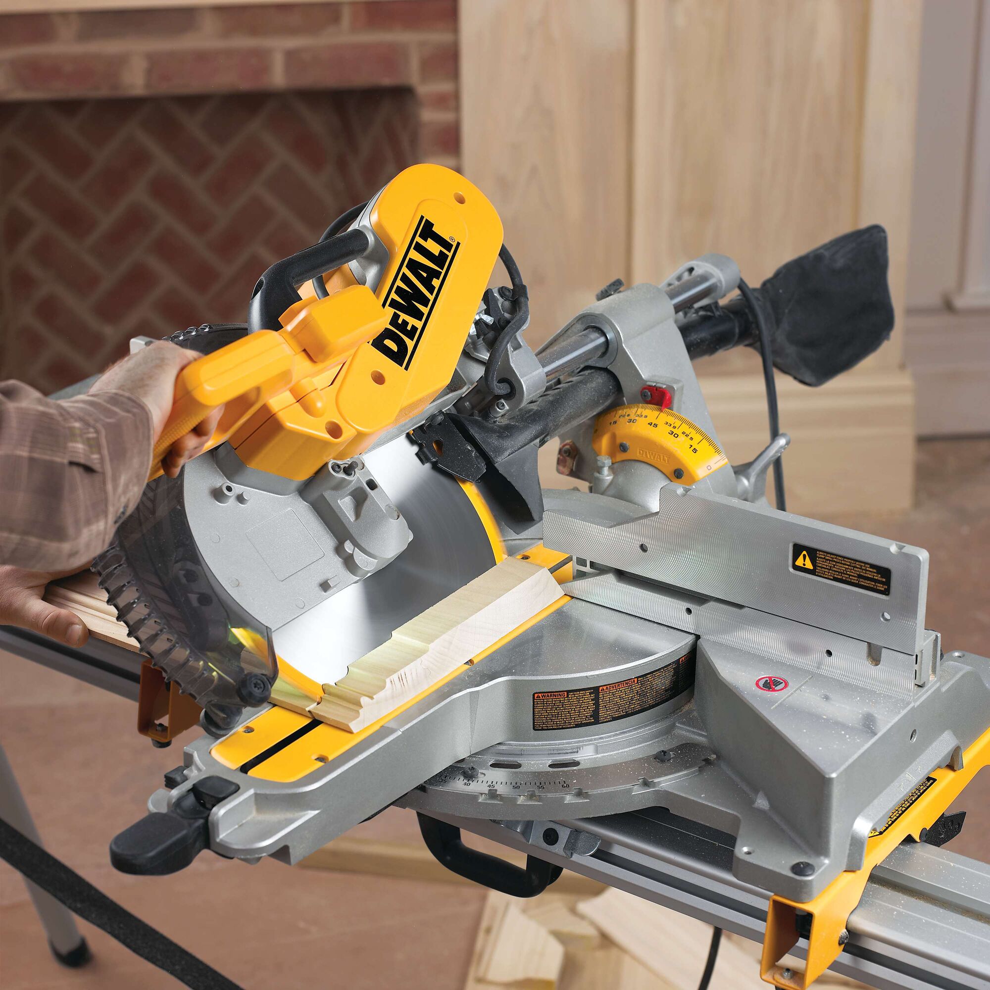 A person operating a DEWALT DWS780 miter saw, cutting a piece of wood. The saw is yellow and gray, and is situated on a workbench indoors.